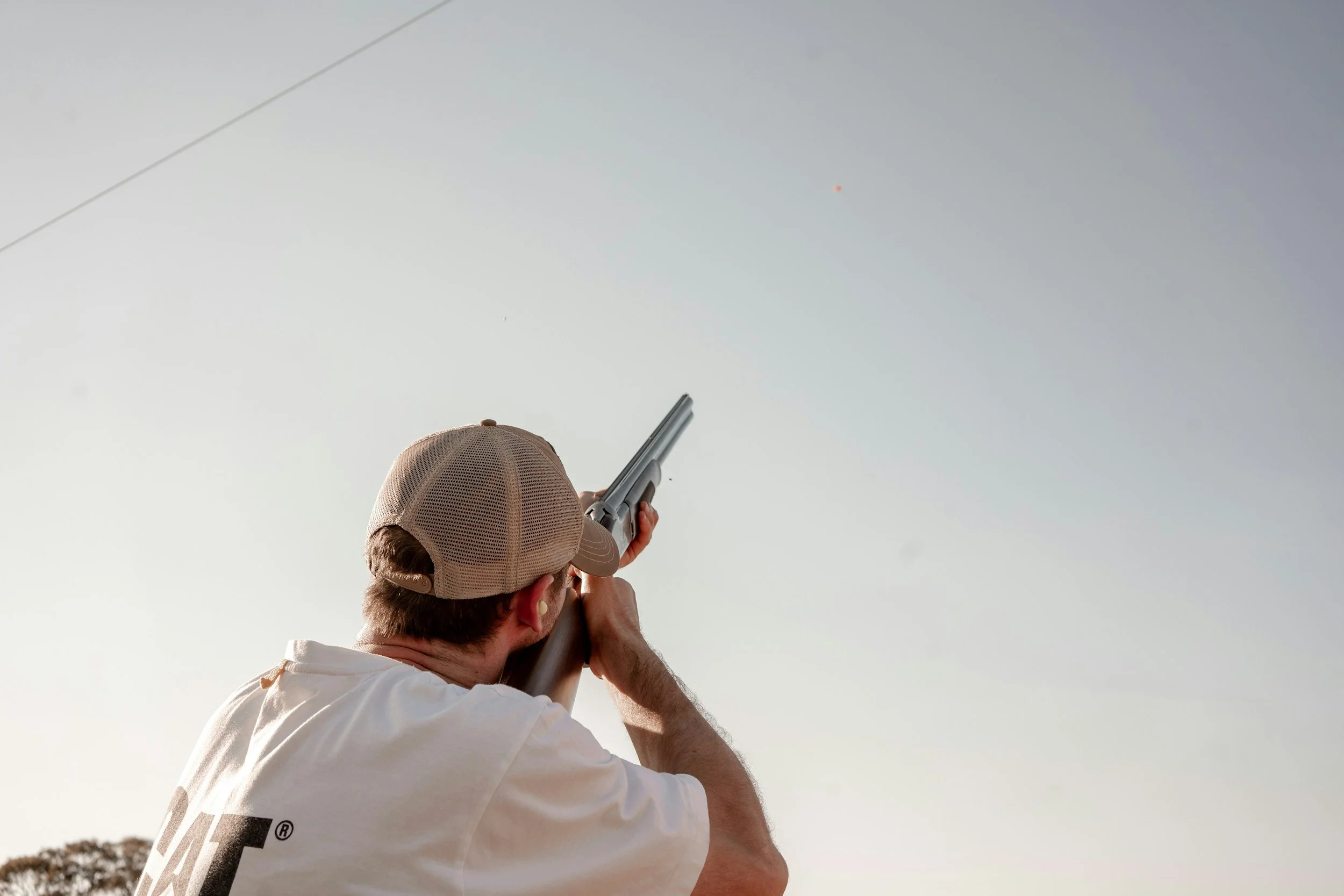 Man wearing beige cap aiming a shotgun towards the sky during daytime.