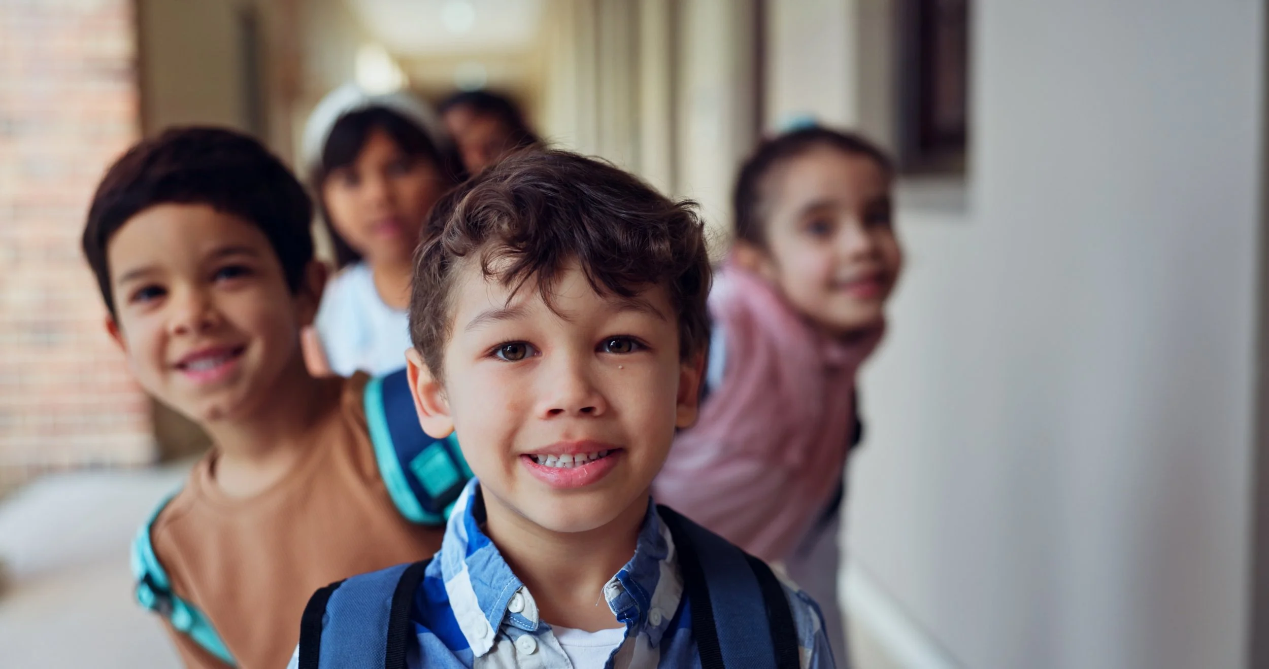 A group of five young children standing in a line, smiling, in a school hallway with windows and brick wall in the background.