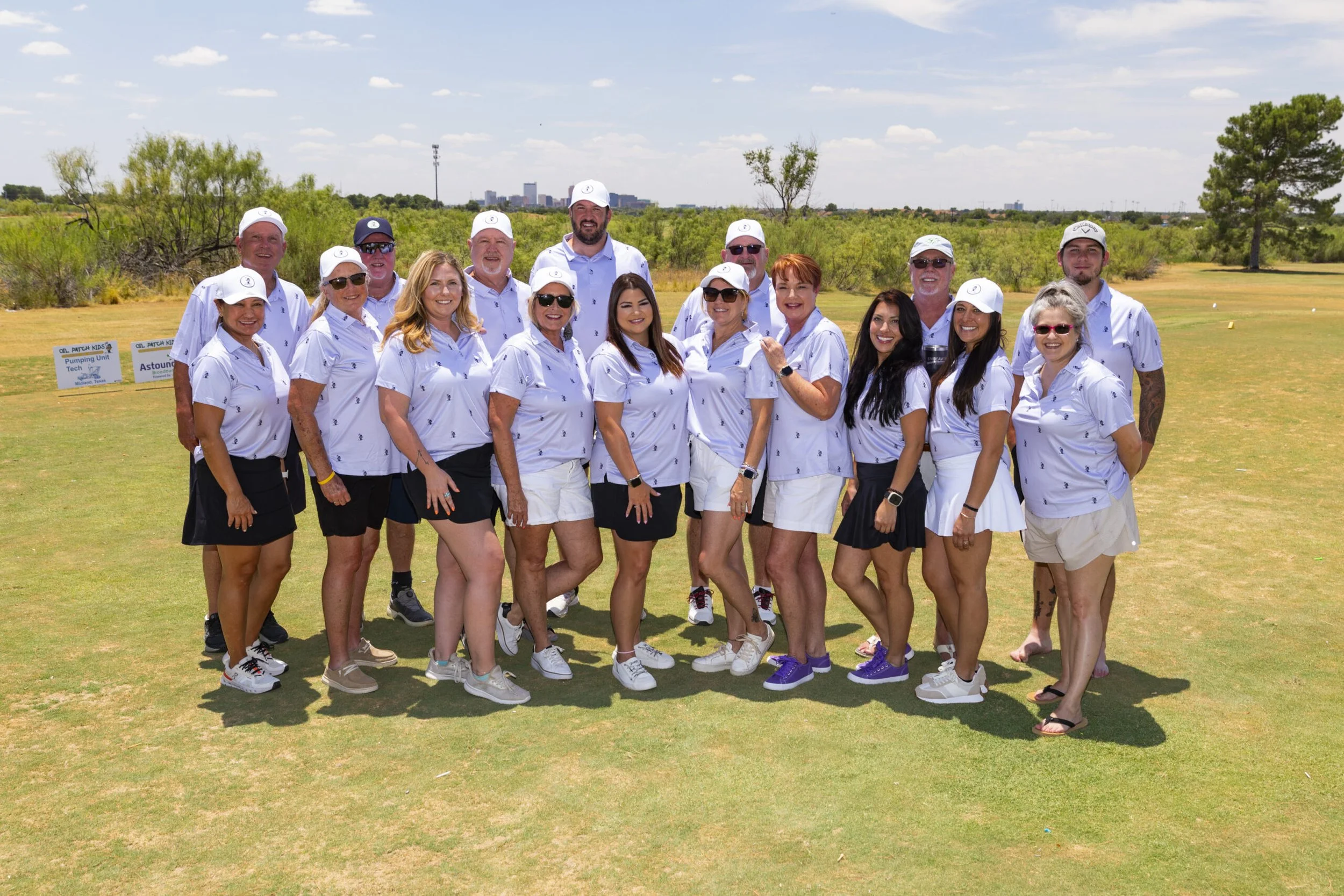 Group of people at a golf course, wearing white shirts and hats, posing for a photo on a sunny day.