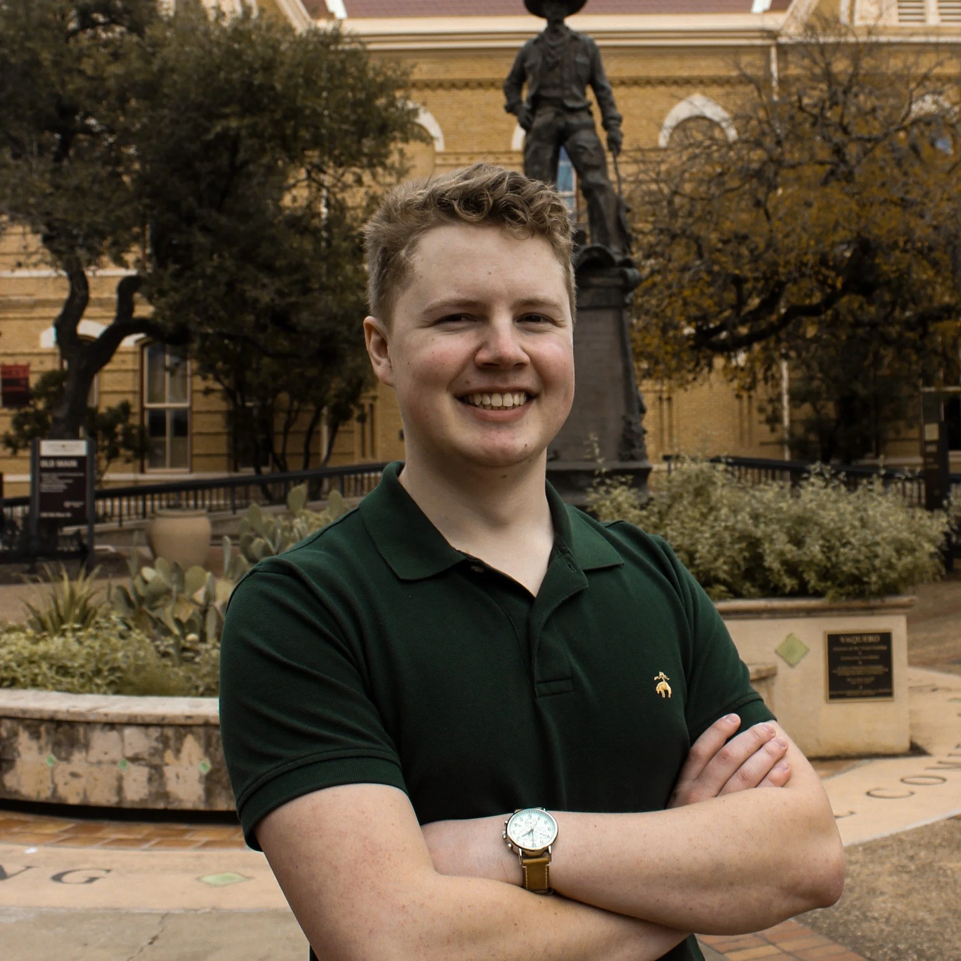 A young man with short, curly hair smiling and standing with arms crossed in front of a historical building. Behind him is a statue of a man in historic attire on a pedestal, surrounded by trees and plants in a garden area.