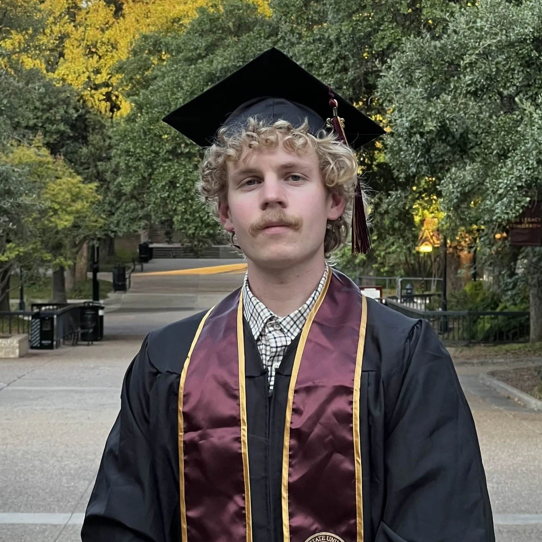 A young man with curly blonde hair and a mustache wearing a black graduation gown and cap, standing outdoors on a pathway with trees in the background.