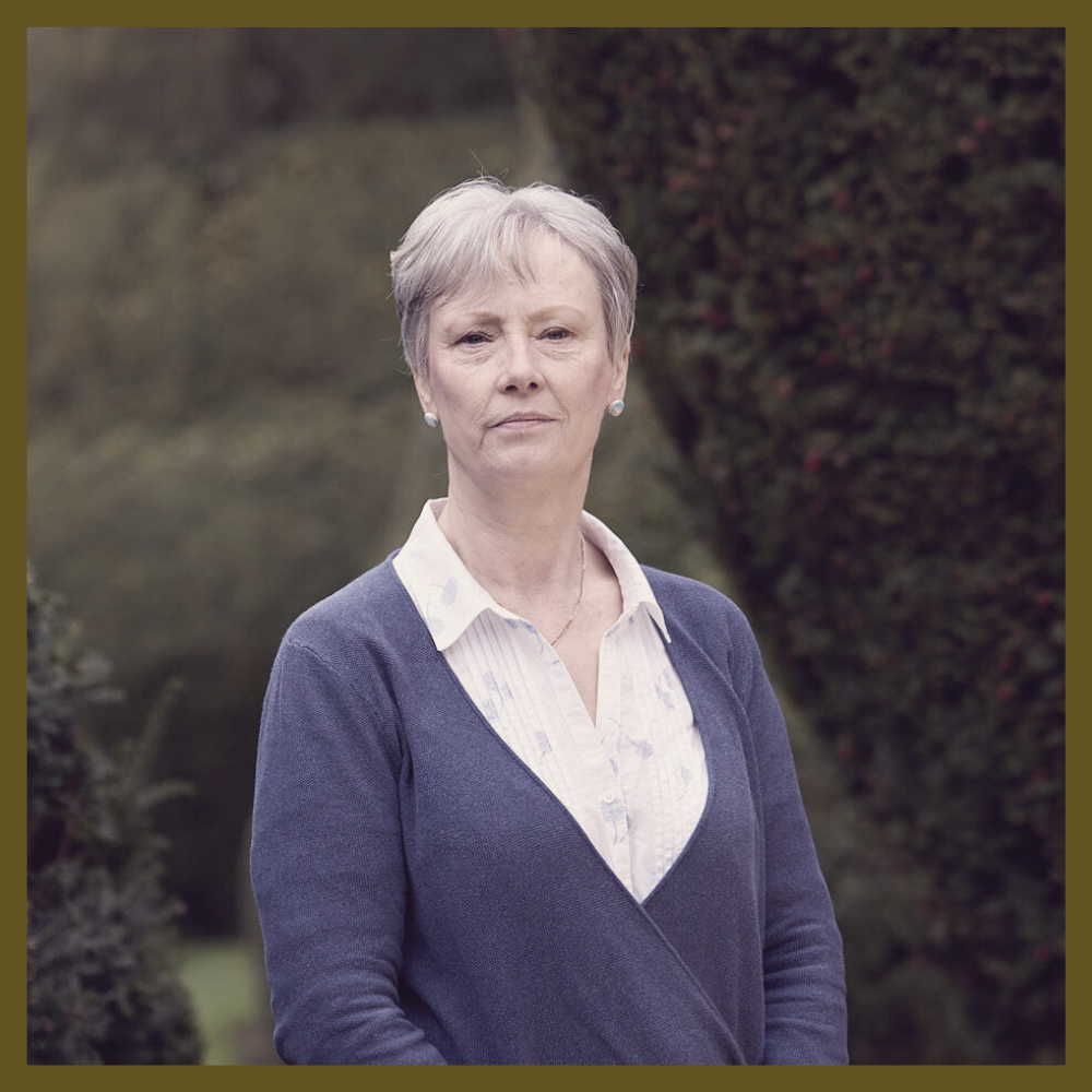 Portrait of a woman with short gray hair, wearing a light-colored blouse and a navy blue cardigan, standing outdoors with blurred trees in the background.