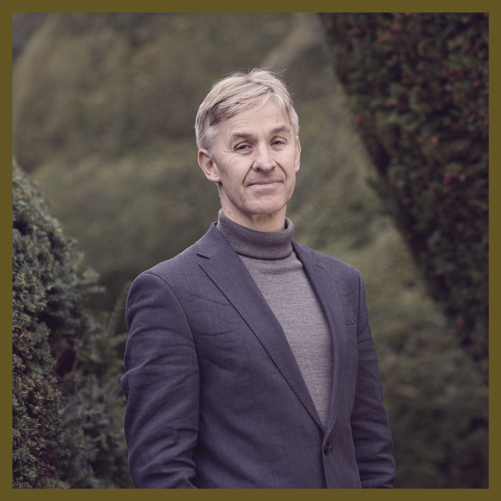 Photograph of a man with short blonde hair, wearing a grey polo neck and a navy blue suit, with greenery in the background. 