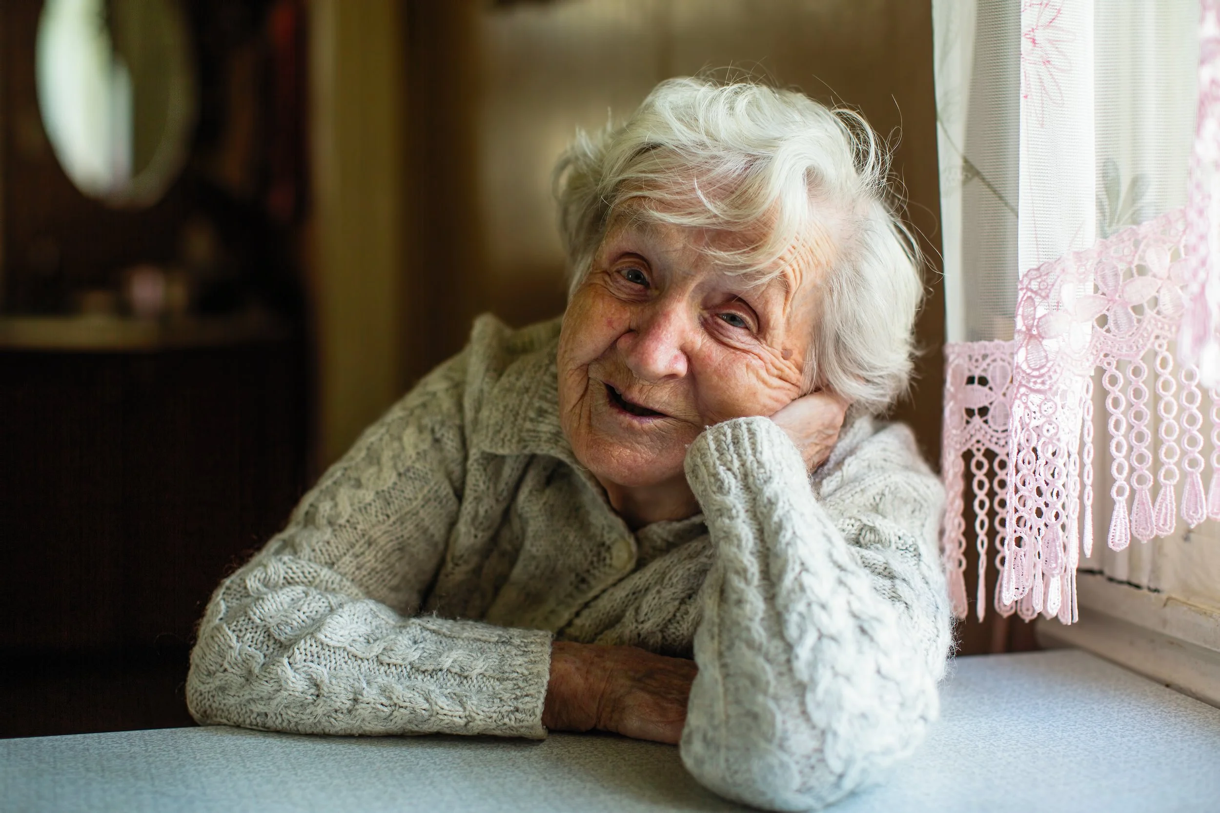 An image of a smiling older woman with short white hair, wearing an oatmeal coloured cardigan and sitting by a window.