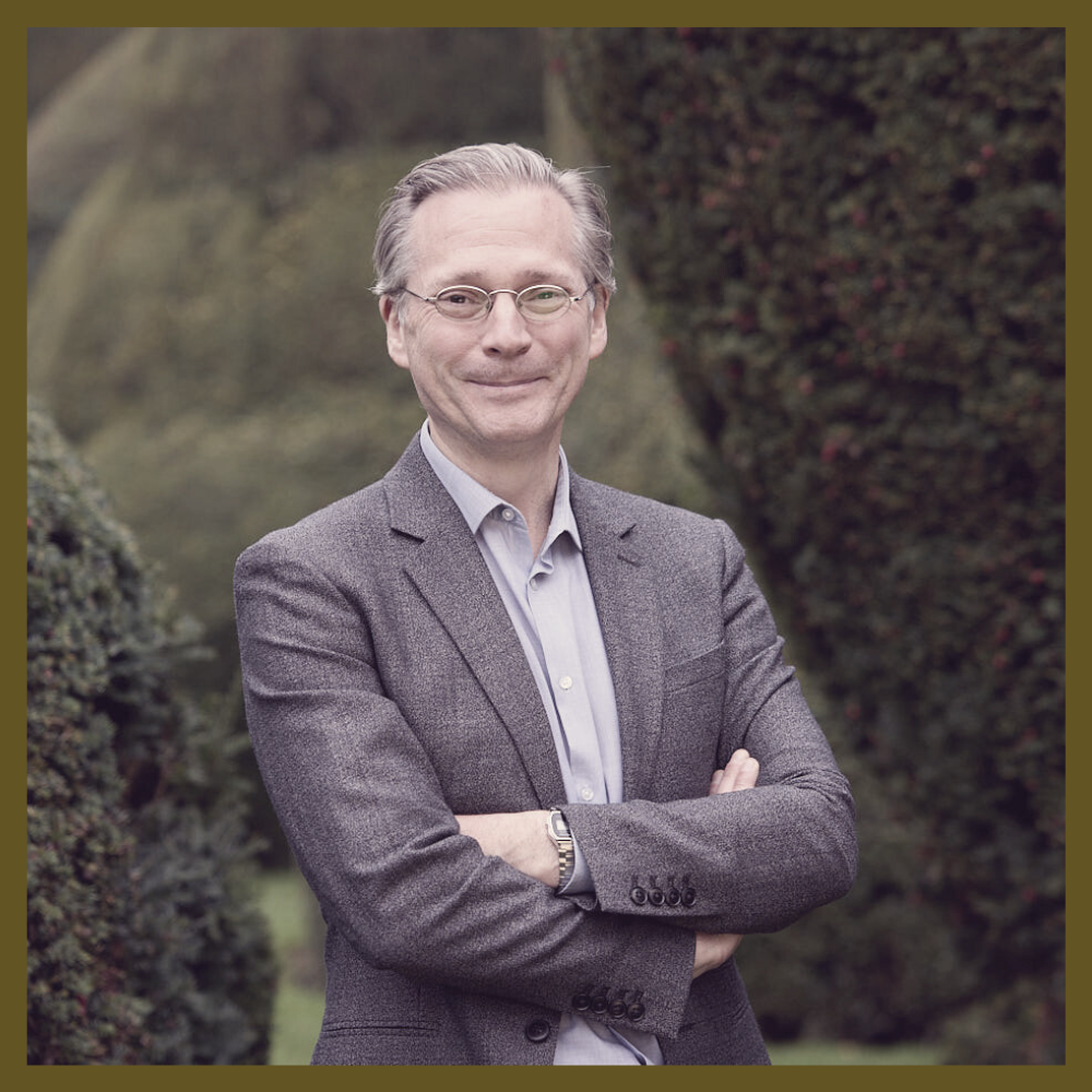 A man with gray hair and glasses, wearing a gray blazer and a light purple button-up shirt, stands outdoors with arms crossed, smiling, with green shrubs and trees in the background.