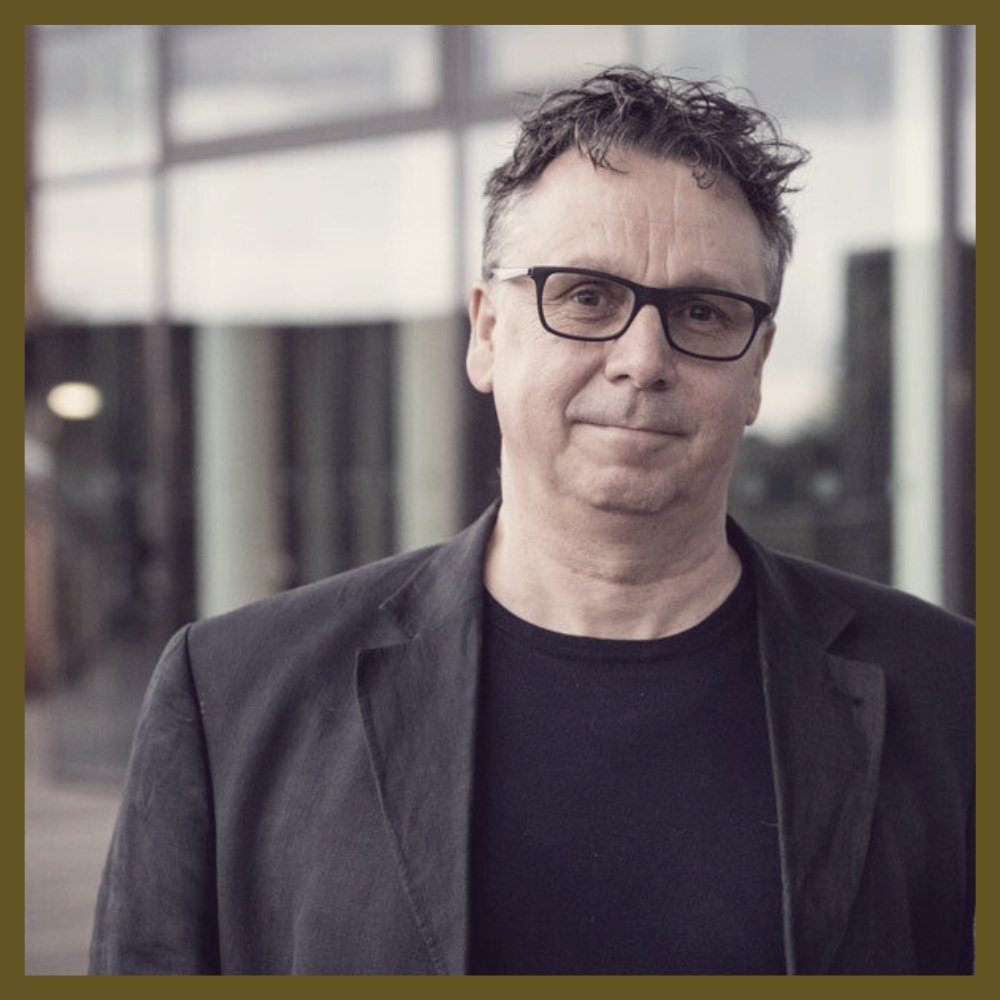 A man with glasses and curly hair, wearing a black jacket and T-shirt, standing outdoors near a modern building.