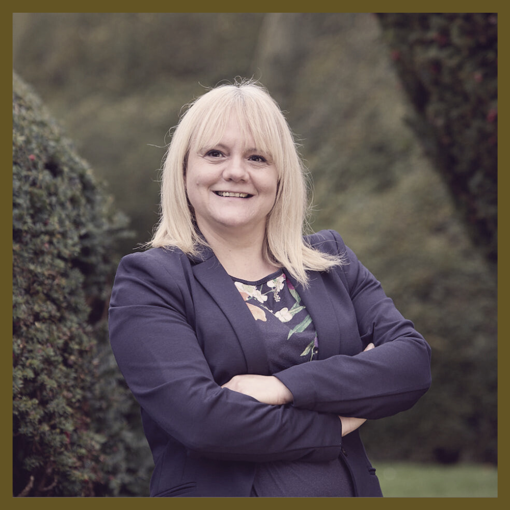 A smiling woman with blonde hair, wearing a dark blazer over a floral top, standing outdoors with her arms crossed, surrounded by greenery in the background.
