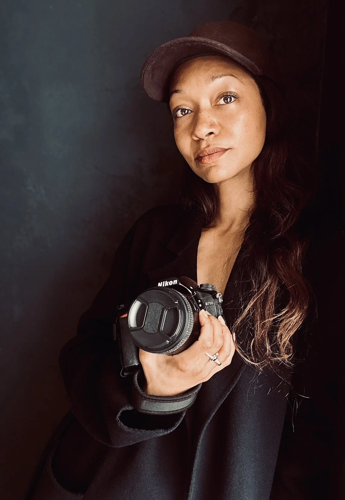 A woman with long dark hair wearing a brown hat and a black coat, holding a Nikon camera, standing against a dark background.