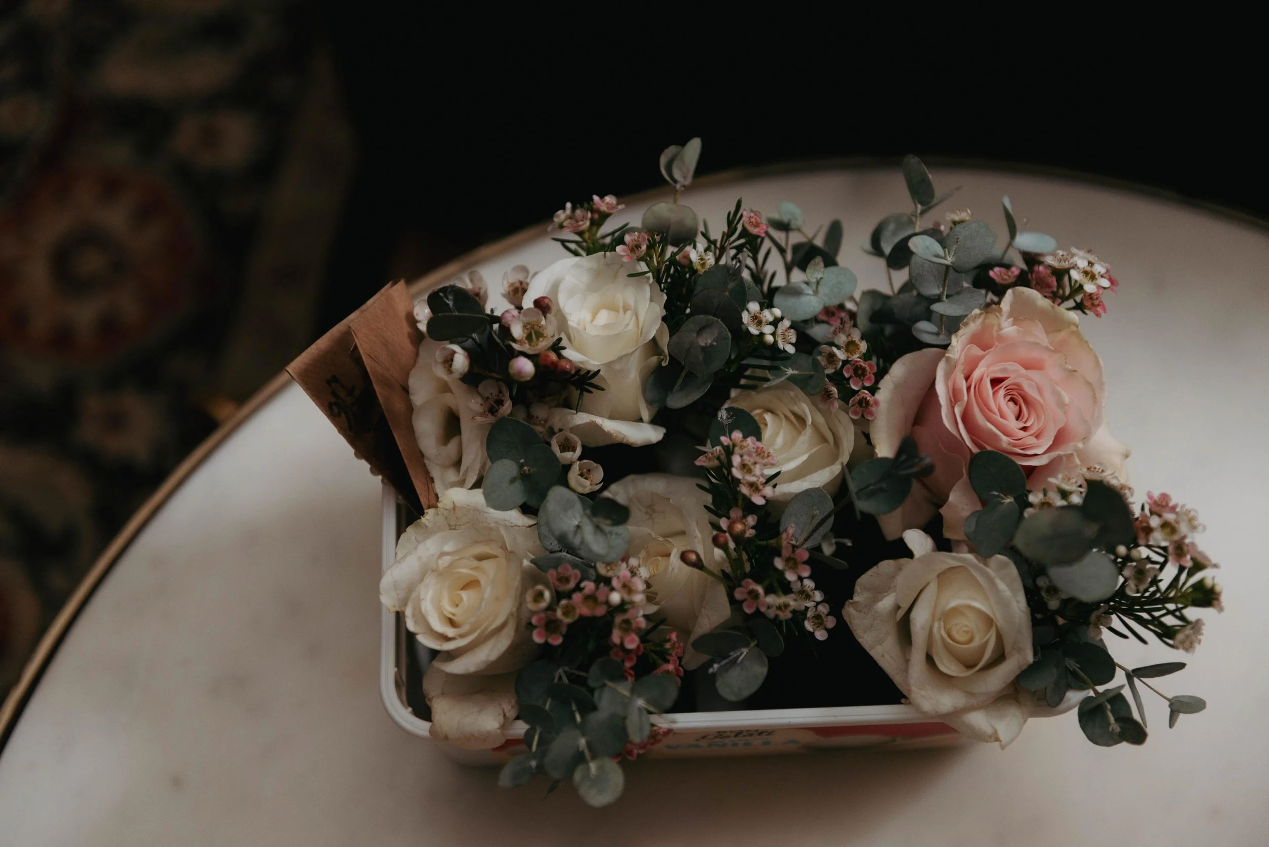 A floral arrangement with white roses, pink roses, small pink and white flowers, and eucalyptus leaves in a small container resting on a white surface.