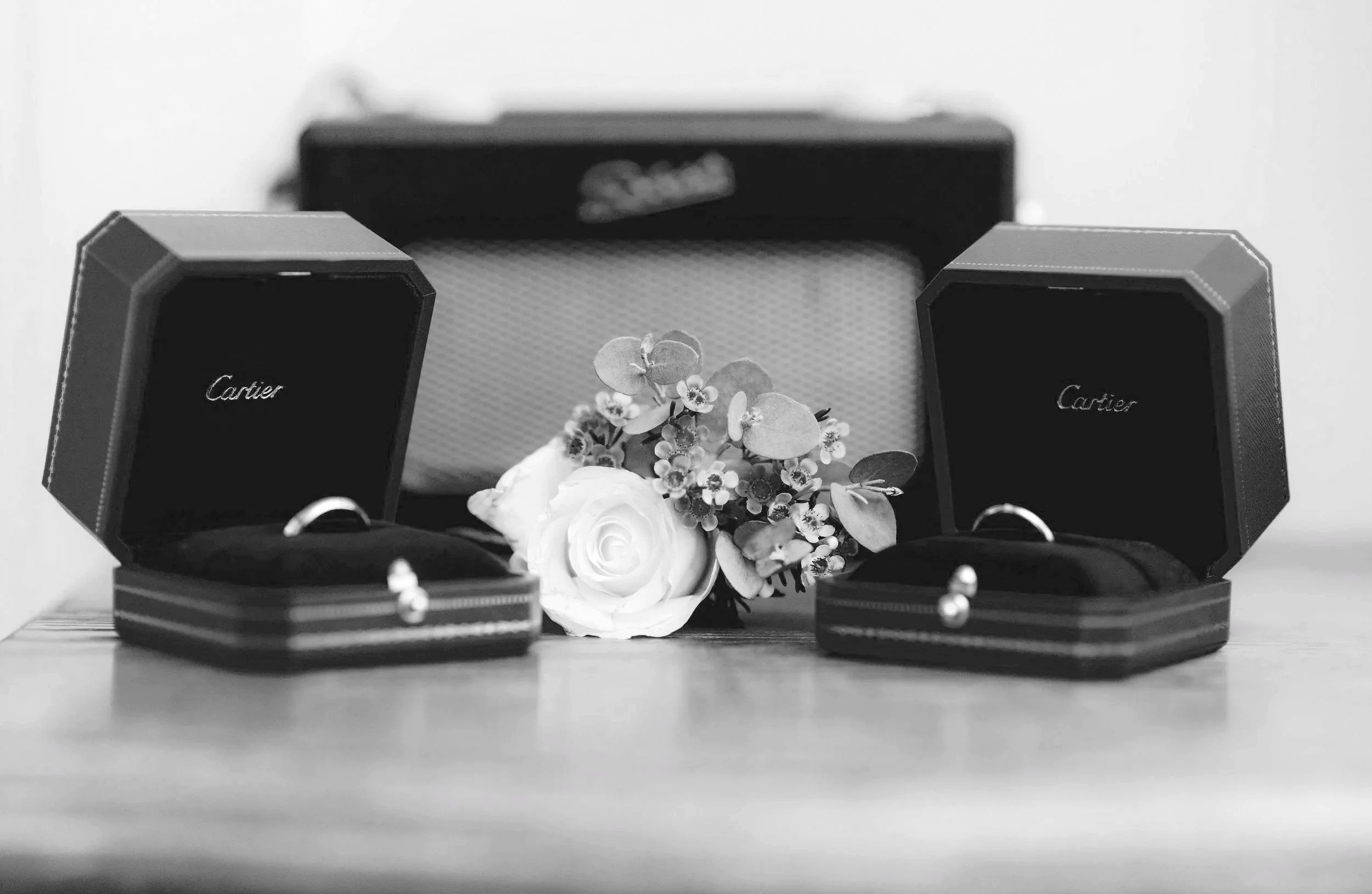 Two open Cartier jewelry boxes with rings inside, a white rose and small flowers in between, and a larger box in the background.