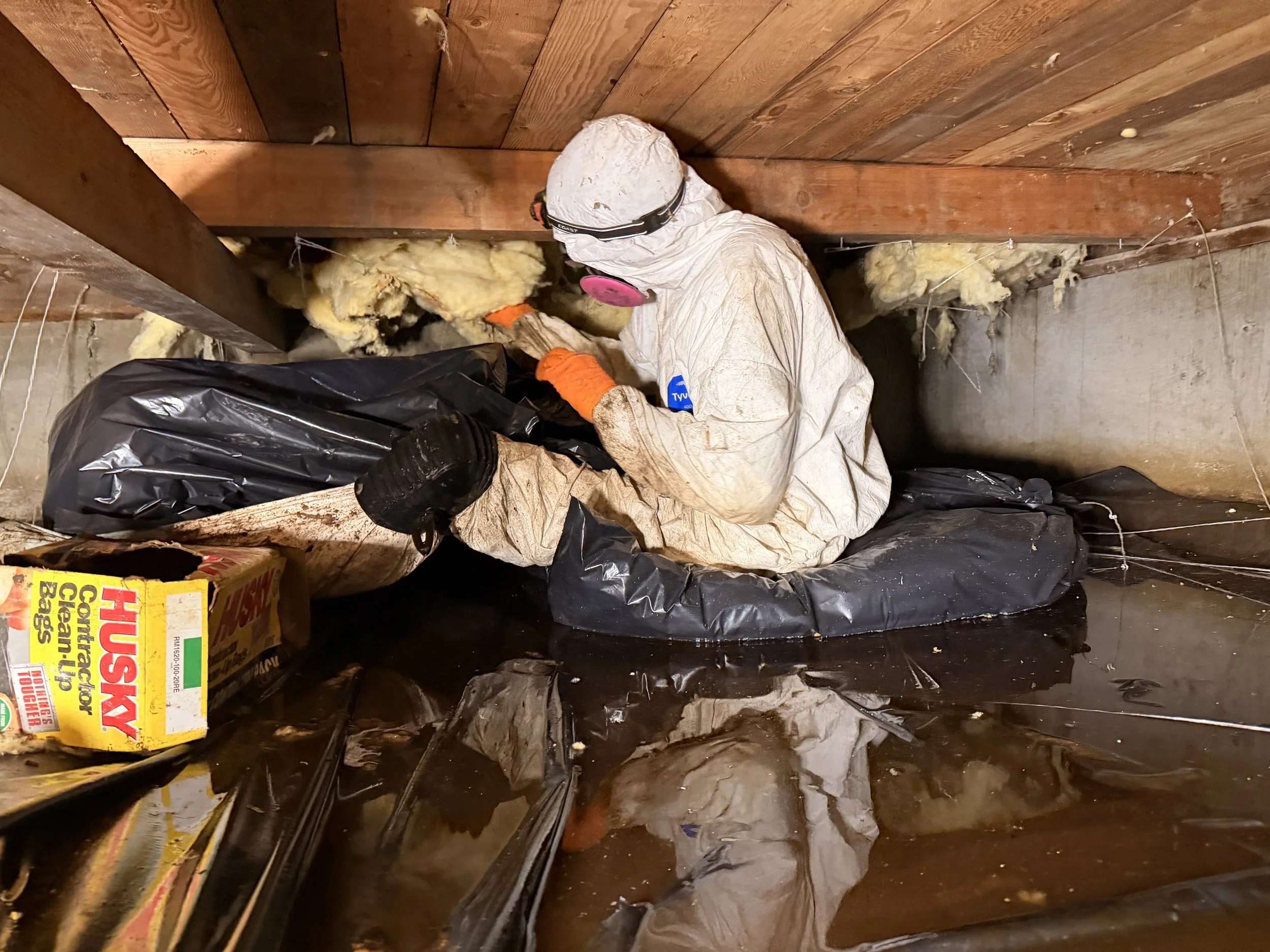 A person wearing protective gear, including a mask and gloves, kneeling in a cramped space, working with insulation material near the floor and wall.