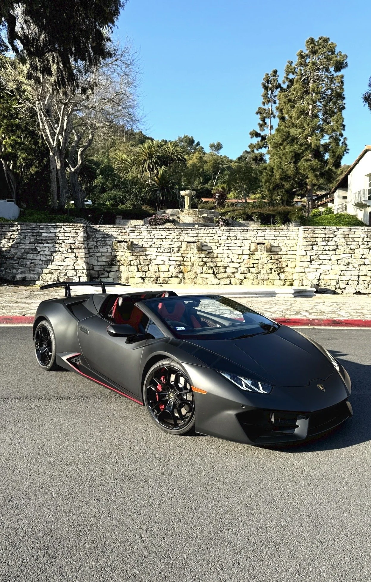 A matte gray Lamborghini Huracan convertible parked on a street with a stone wall and trees in the background.