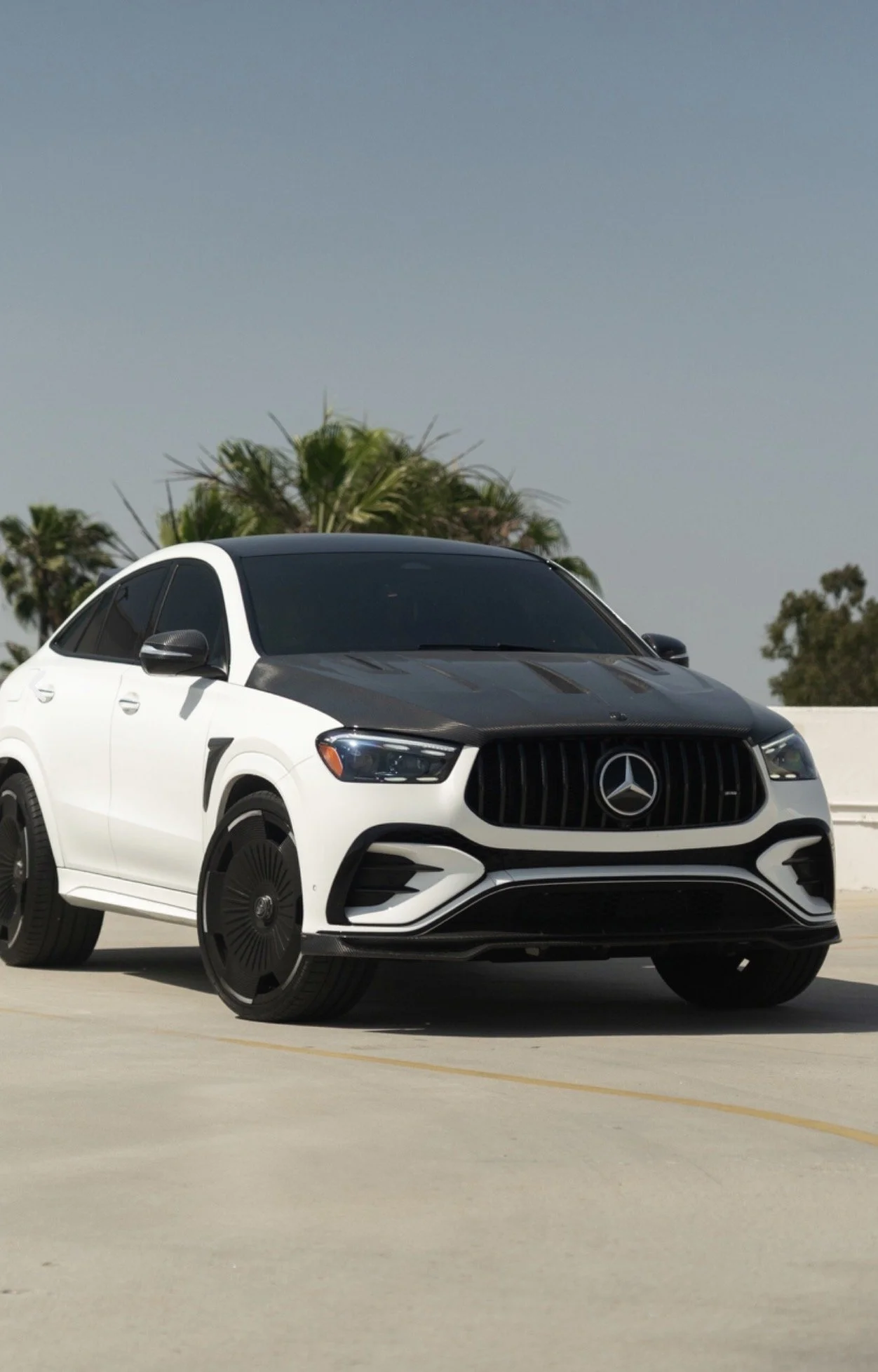 A white Mercedes-Benz sedan with a black hood and black wheel covers parked outdoors under a clear sky with palm trees in the background.
