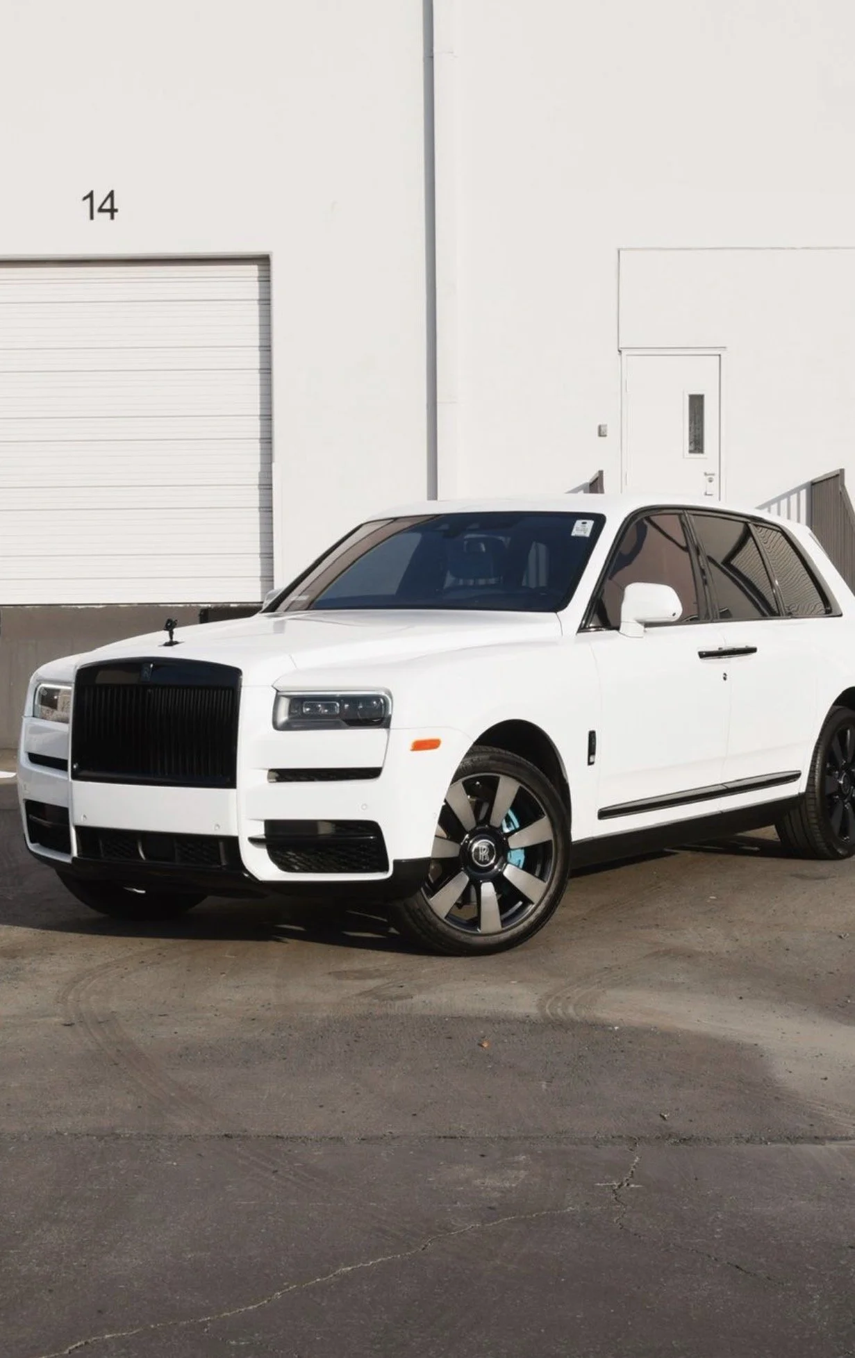 A white Rolls-Royce SUV parked on a concrete surface in front of a white industrial building with a garage door and a side door.