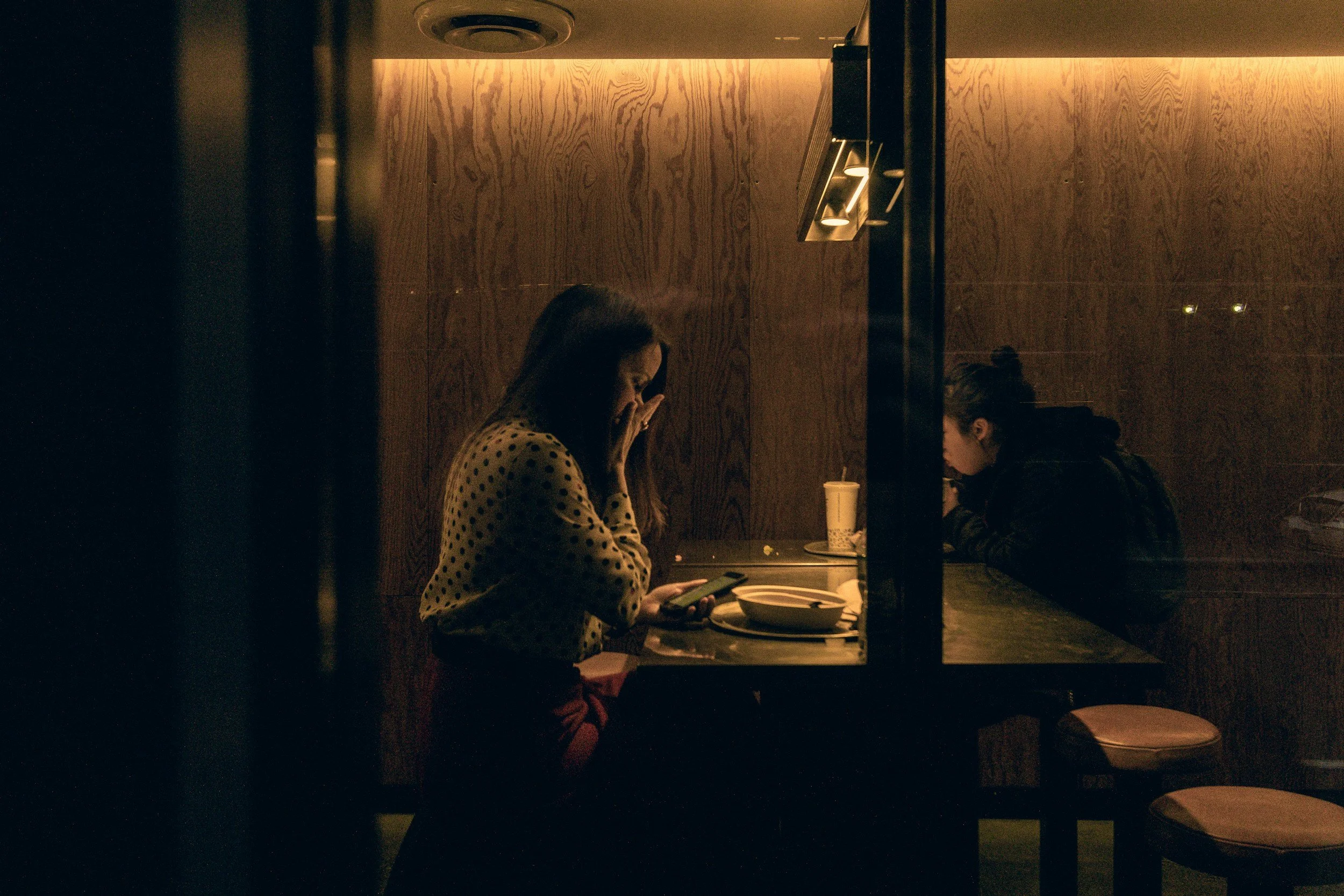 Two women sitting at a table in a dimly lit restaurant or cafe, one looking at a phone and the other with her hand near her mouth, with drinks on the table.