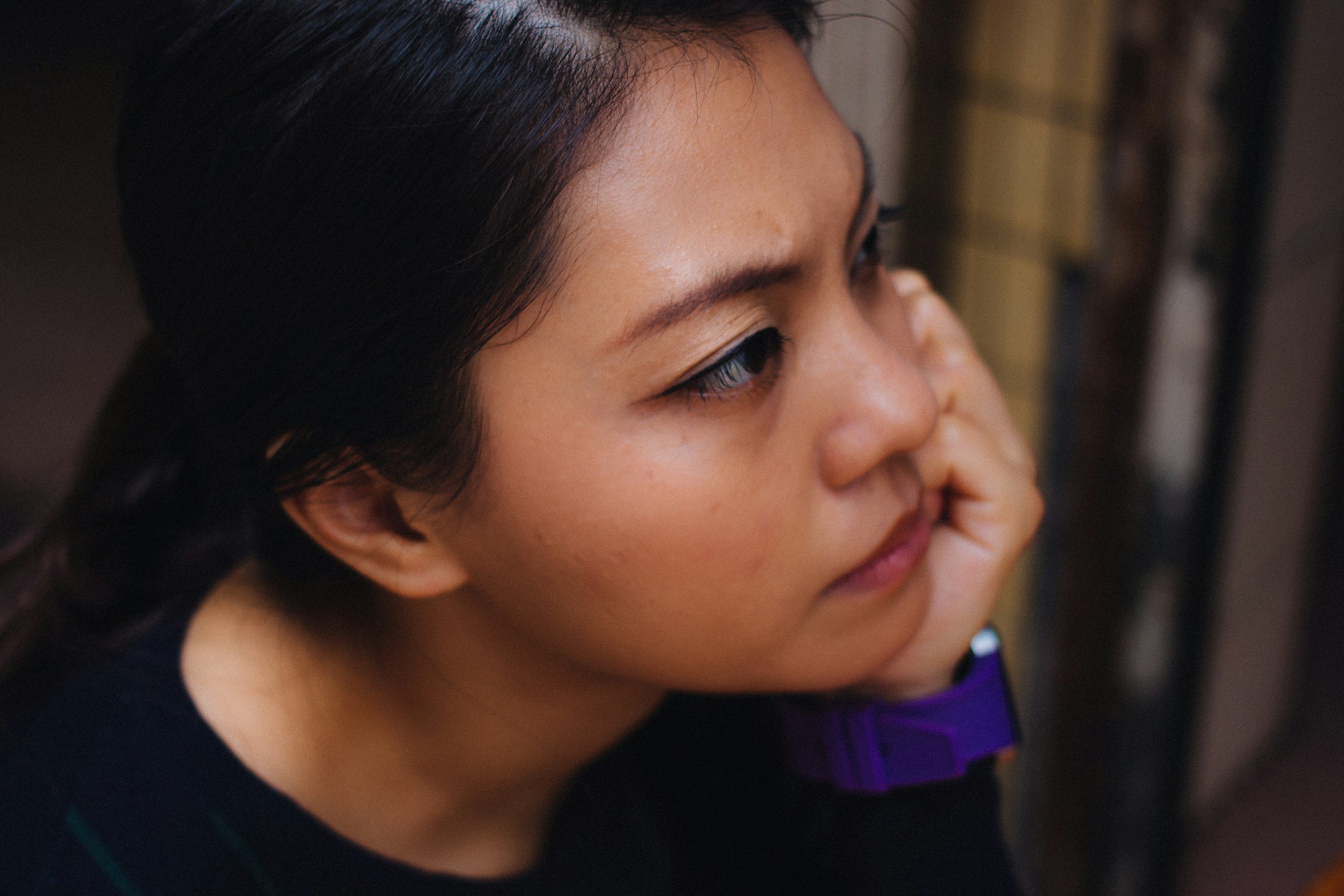 Close-up of a woman with dark hair, touching her face with her hand, wearing a dark top and purple clothing underneath.