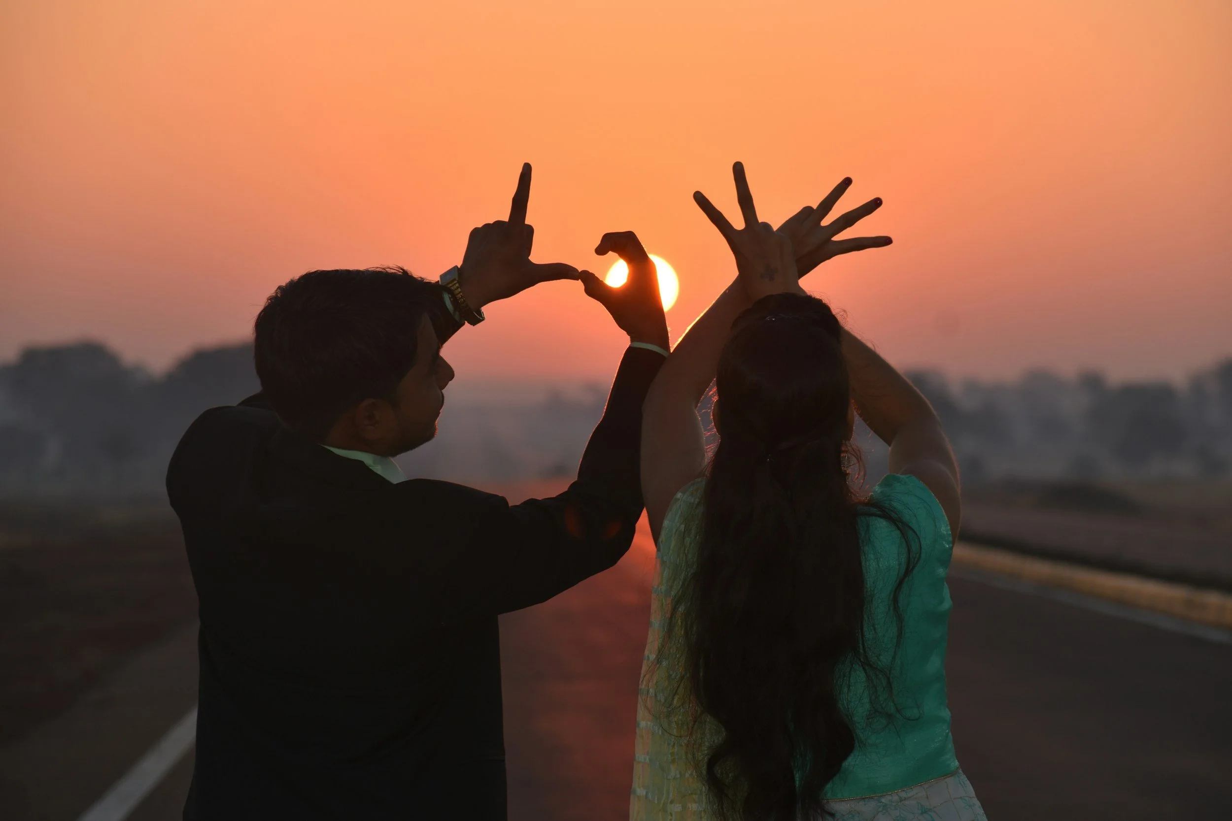 Two people, a man and a woman, standing on a road at sunset, making a heart shape with their hands around the sun.