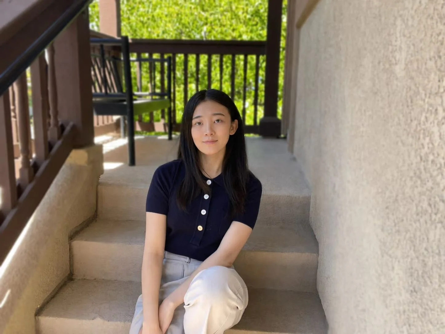 A young woman with black hair wearing a beige cardigan sitting indoors with a plant and window in the background.