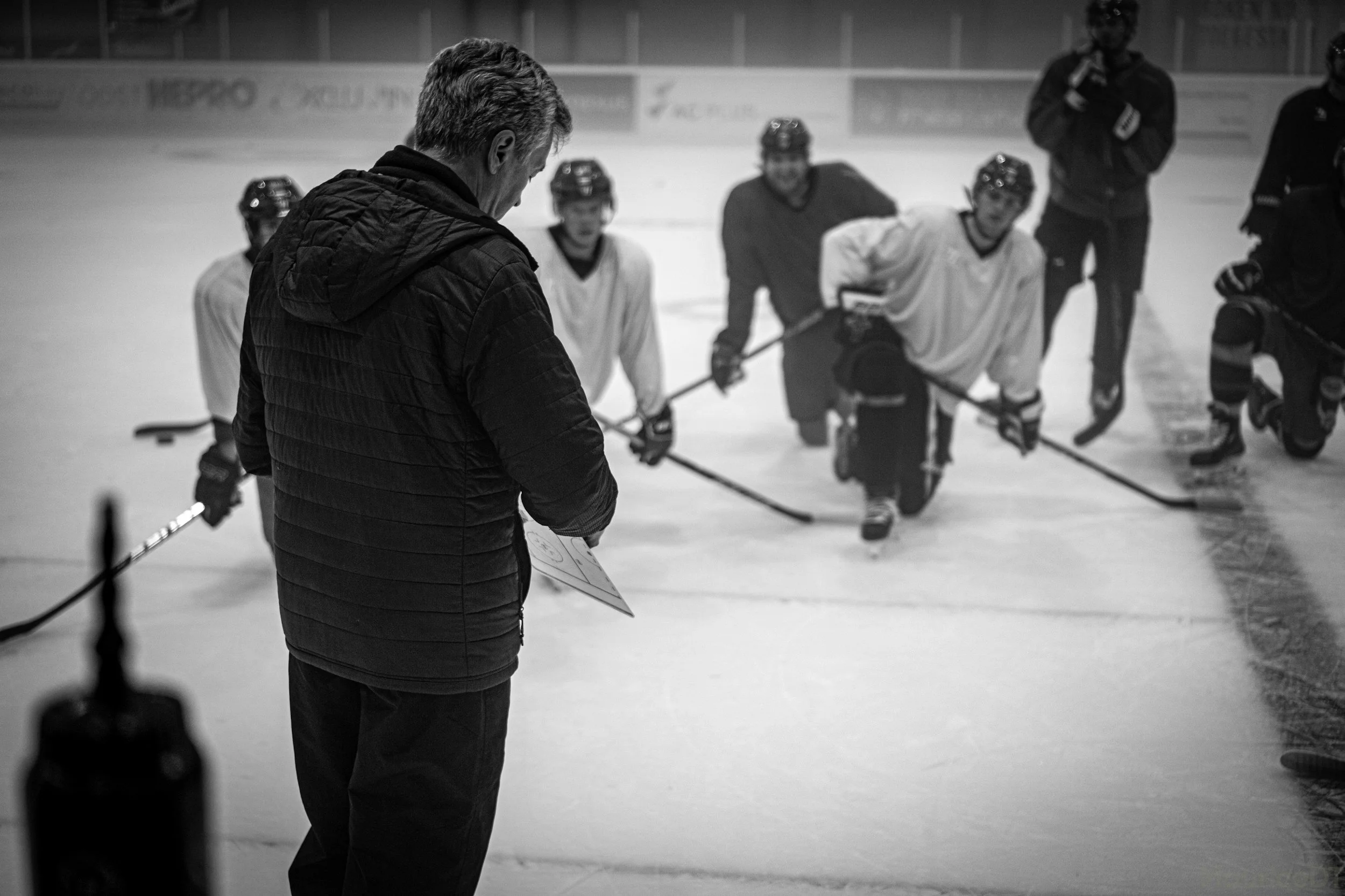 A coach or instructor standing on an ice hockey rink, holding a clipboard and diagram, while coaching a group of five hockey players who are kneeling on the ice, listening attentively.