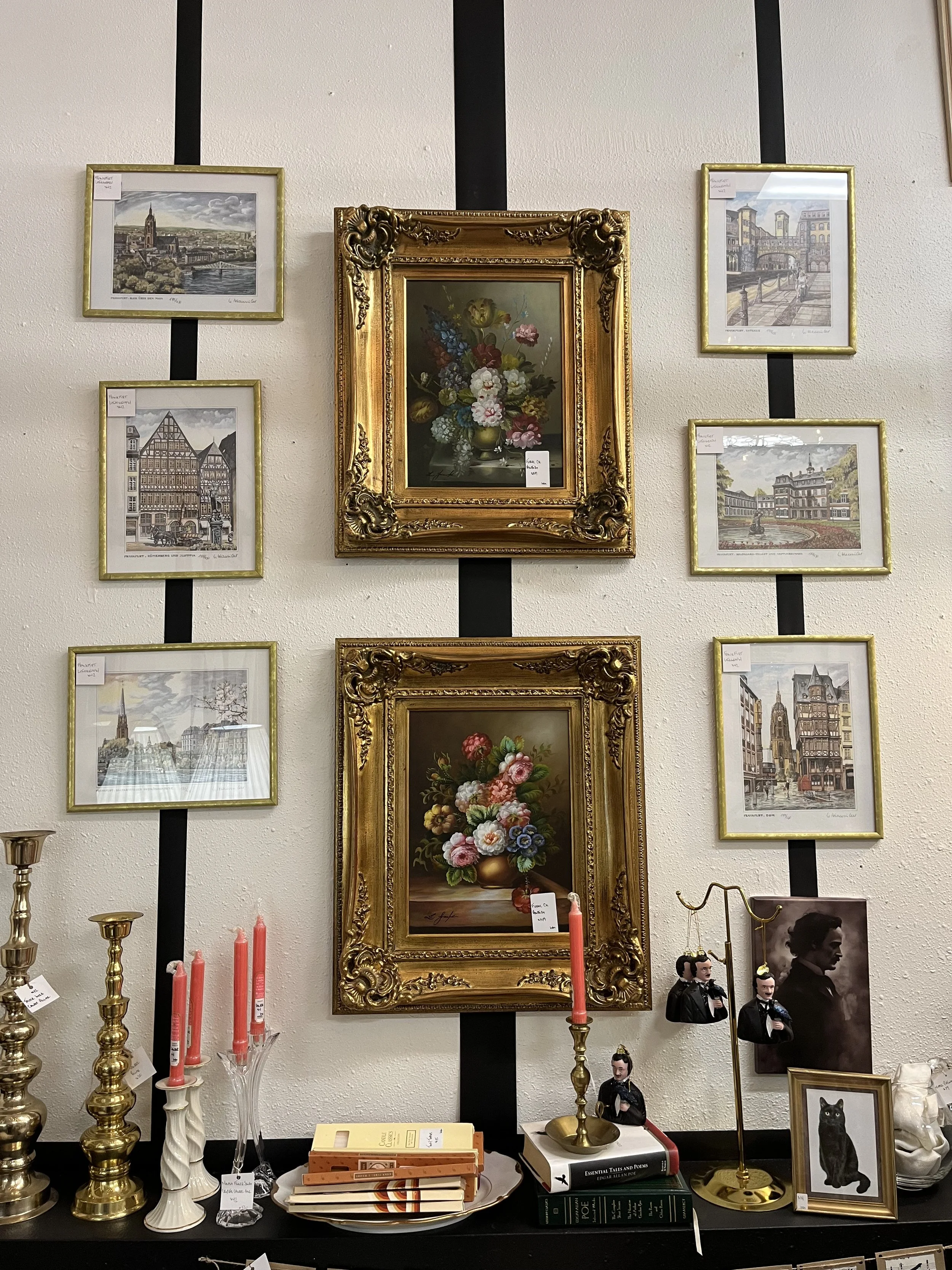 A display of various framed artworks, including paintings and prints, arranged on a wall and shelves. The central piece is an ornate gold-framed still life of flowers, surrounded by pictures of cityscapes, architecture, and a black cat.