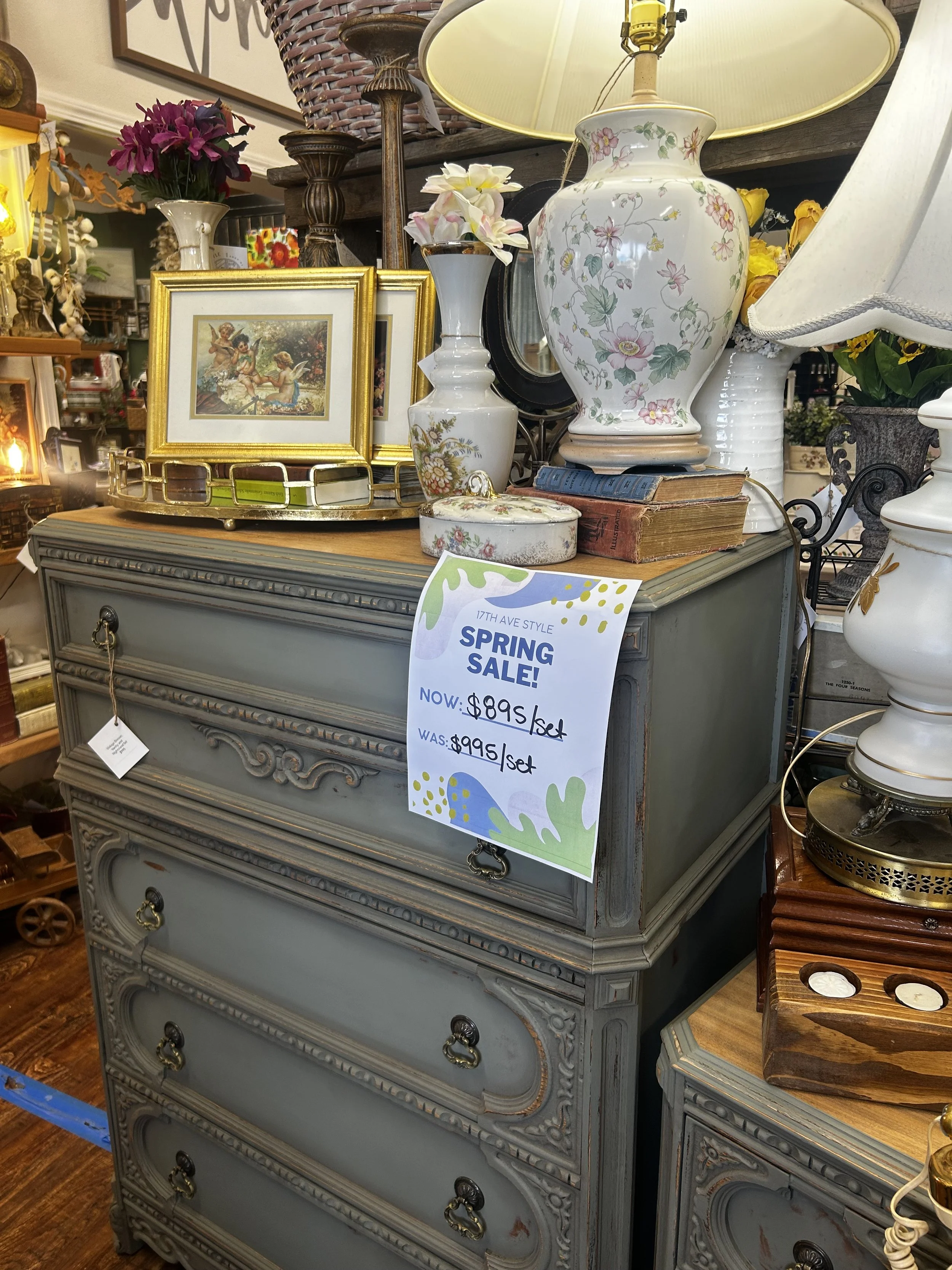 A vintage-style gray dresser with ornate details and several drawers, topped with decorative vases, framed pictures, and a stack of old books. A sign on the dresser indicates a spring sale with prices marked for sets. Several lamps, flowers, and decorative items are visible in a cluttered antique store setting.