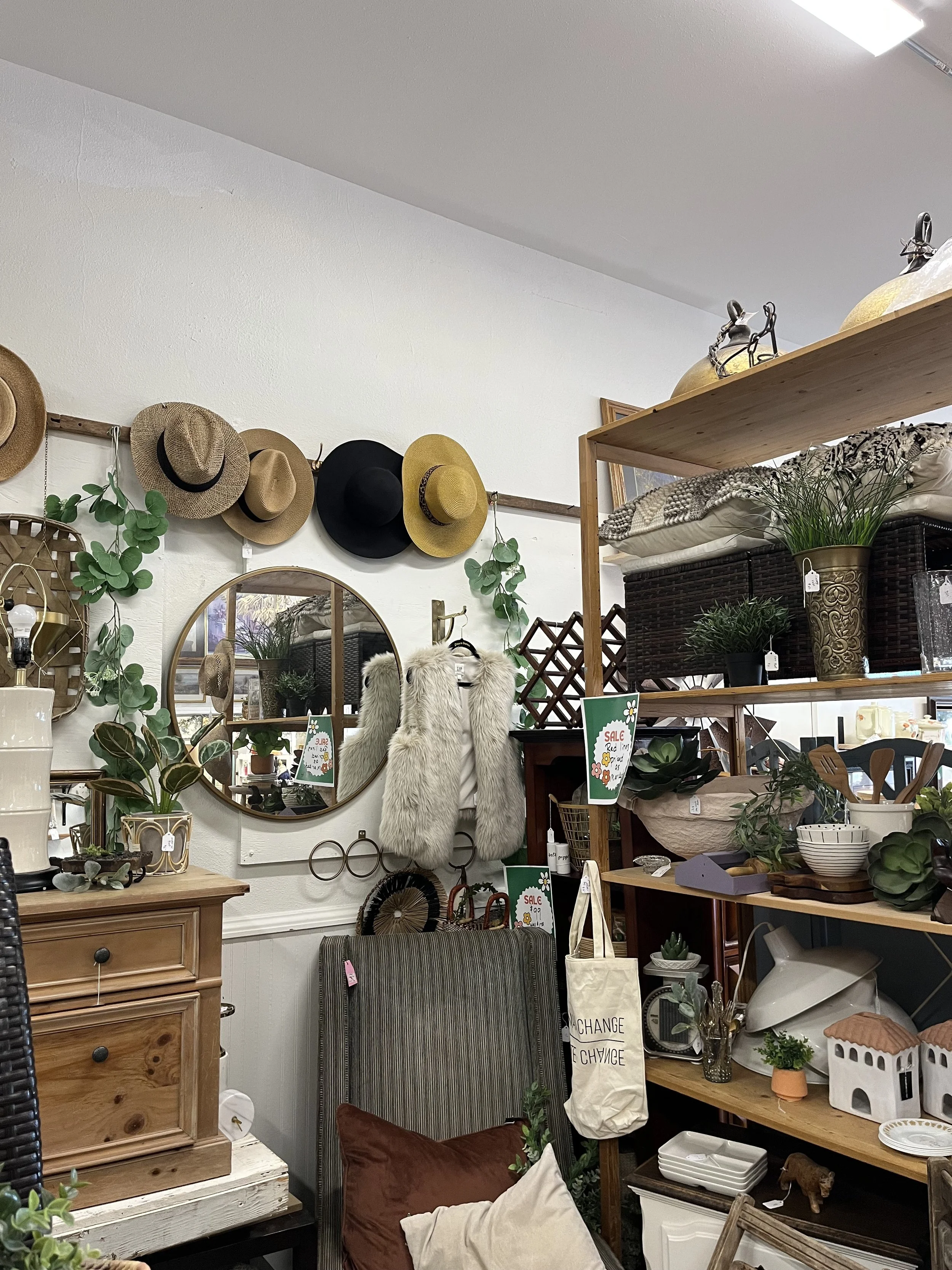 A display of home decor items in a thrift store, including hats on the wall, mirrors, potted plants, pillows, and various small furniture and decorative objects.