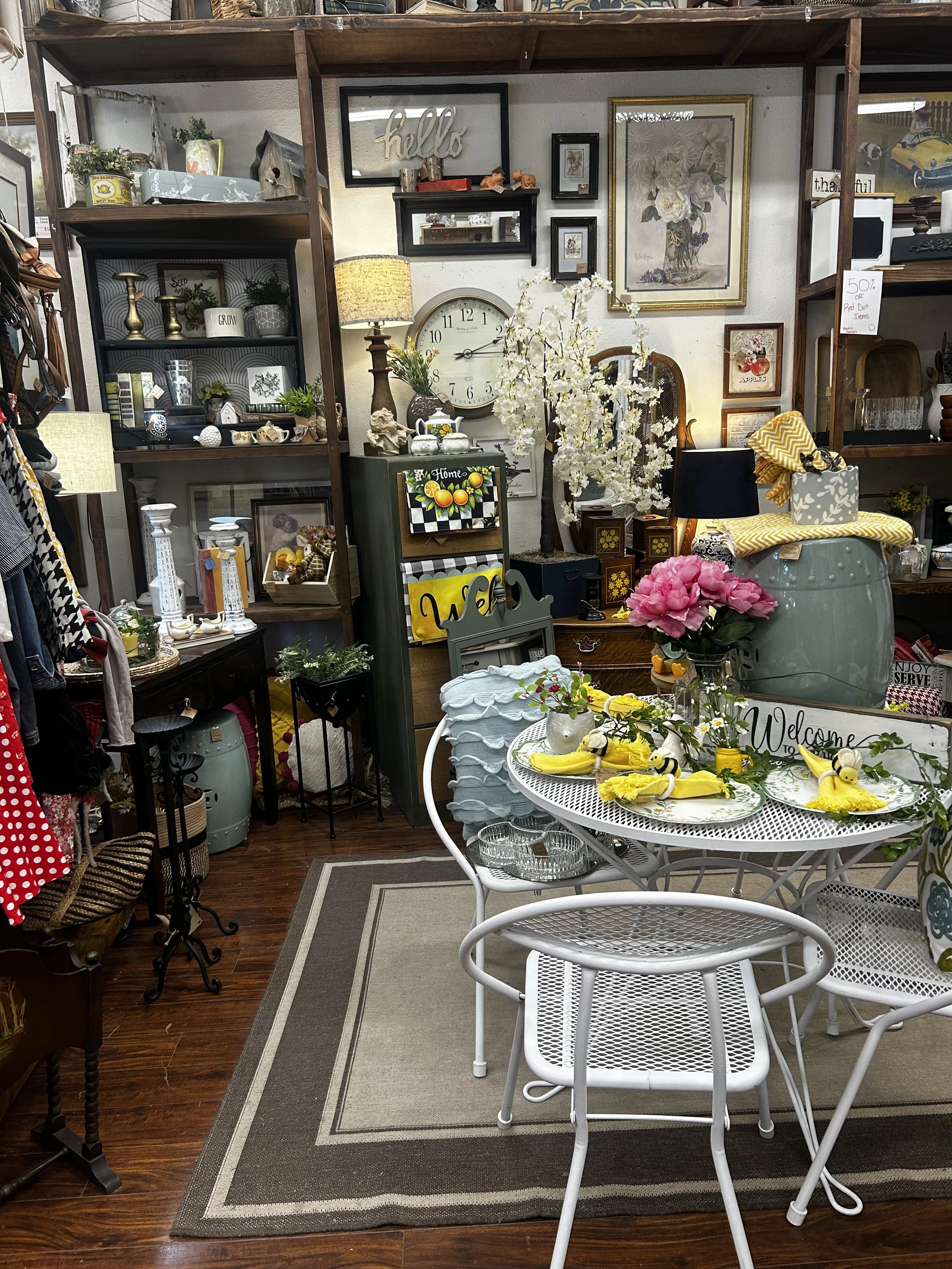 A cozy store display with various home decor items, figurines, a large wall clock, framed artwork, a white round patio table with yellow and bee-themed table settings, a gray and white area rug, and wooden shelving filled with decorative objects and plants.