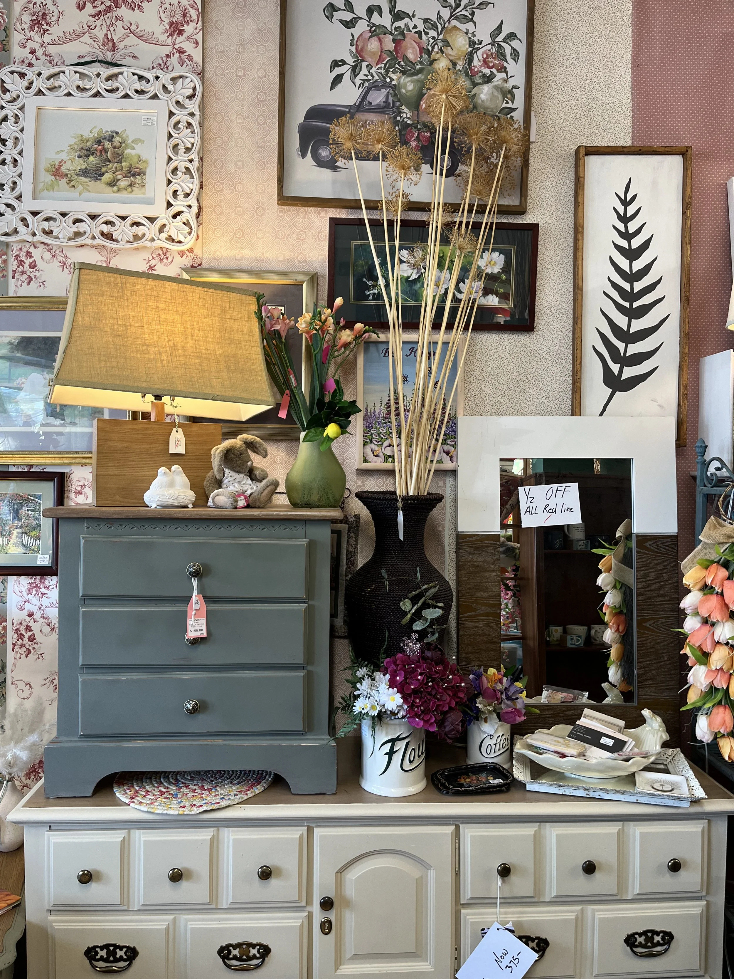 A vintage-style light blue dresser decorated with various flowers, a plush bunny, and small decorative objects. The background features a wall arrangement of framed botanical and floral artwork, a tall black vase with dried plants, and a mirror with a price tag.