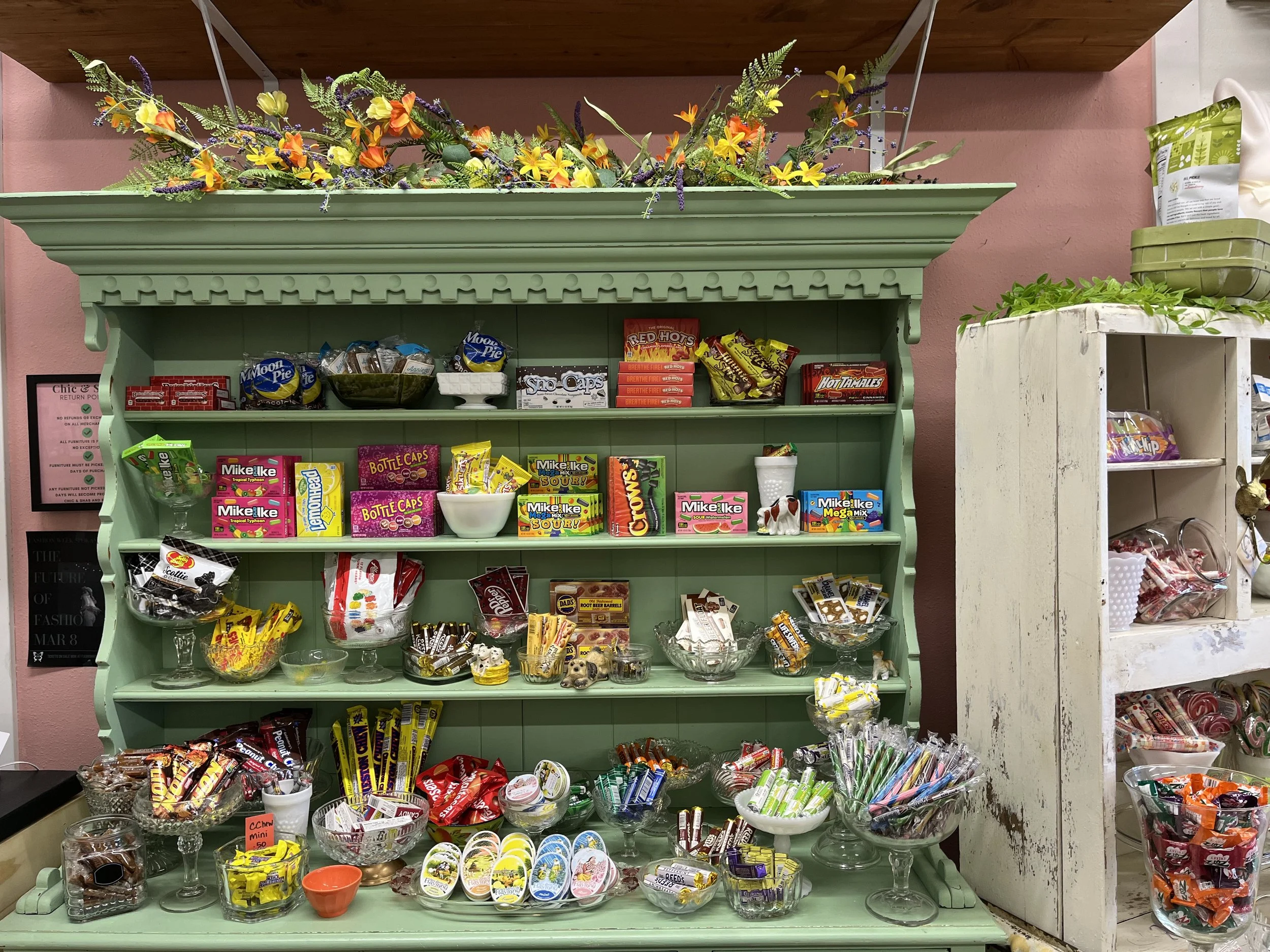 A vintage green candy display shelf filled with various candies and snacks, with decorative flowers on top and pink walls in the background.