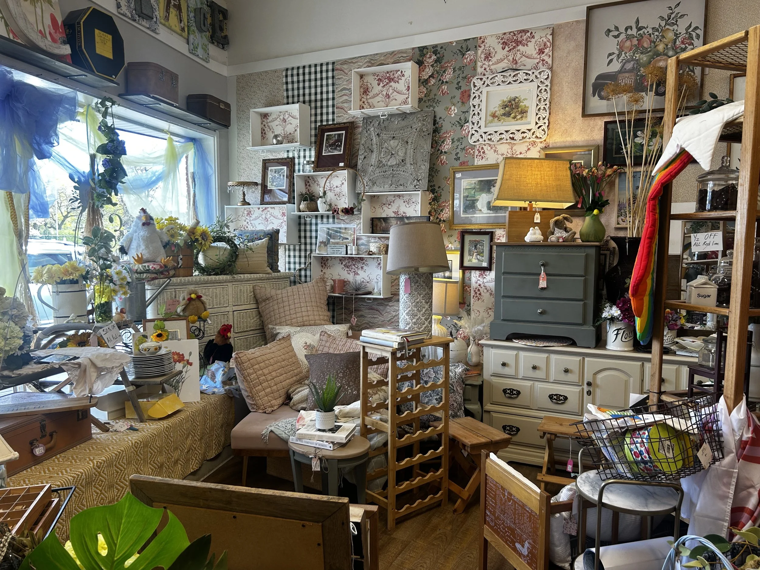Interior of an eclectic store with various framed pictures, floral and checkered wallpaper, shelves with decor items, a table with floral arrangements, and a window with sheer blue and yellow curtains.