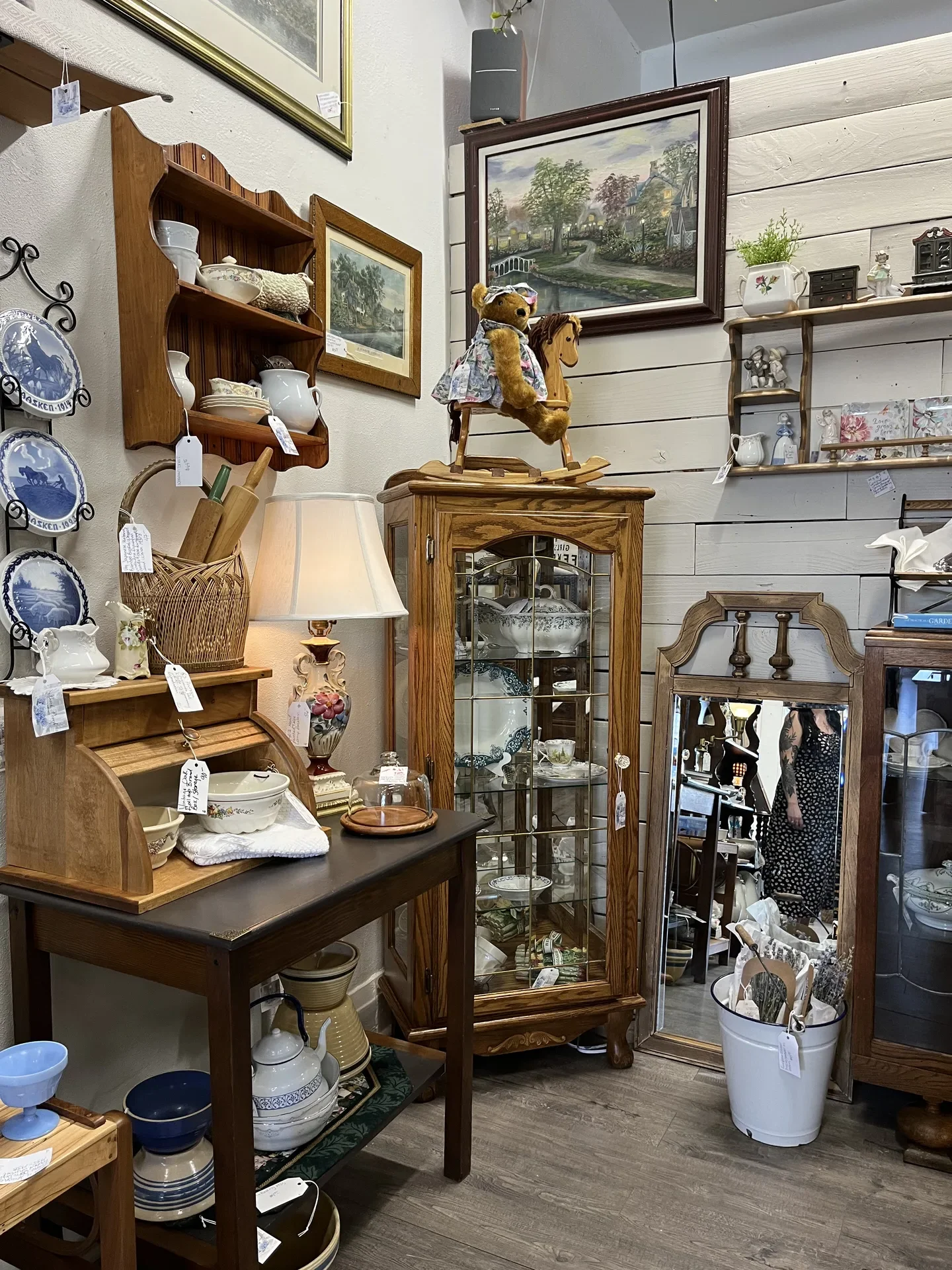Various antique and vintage ceramic and pottery pieces displayed on shelves and tables in a store, with framed landscape paintings on the wall and a teddy bear on a wooden horse toy.