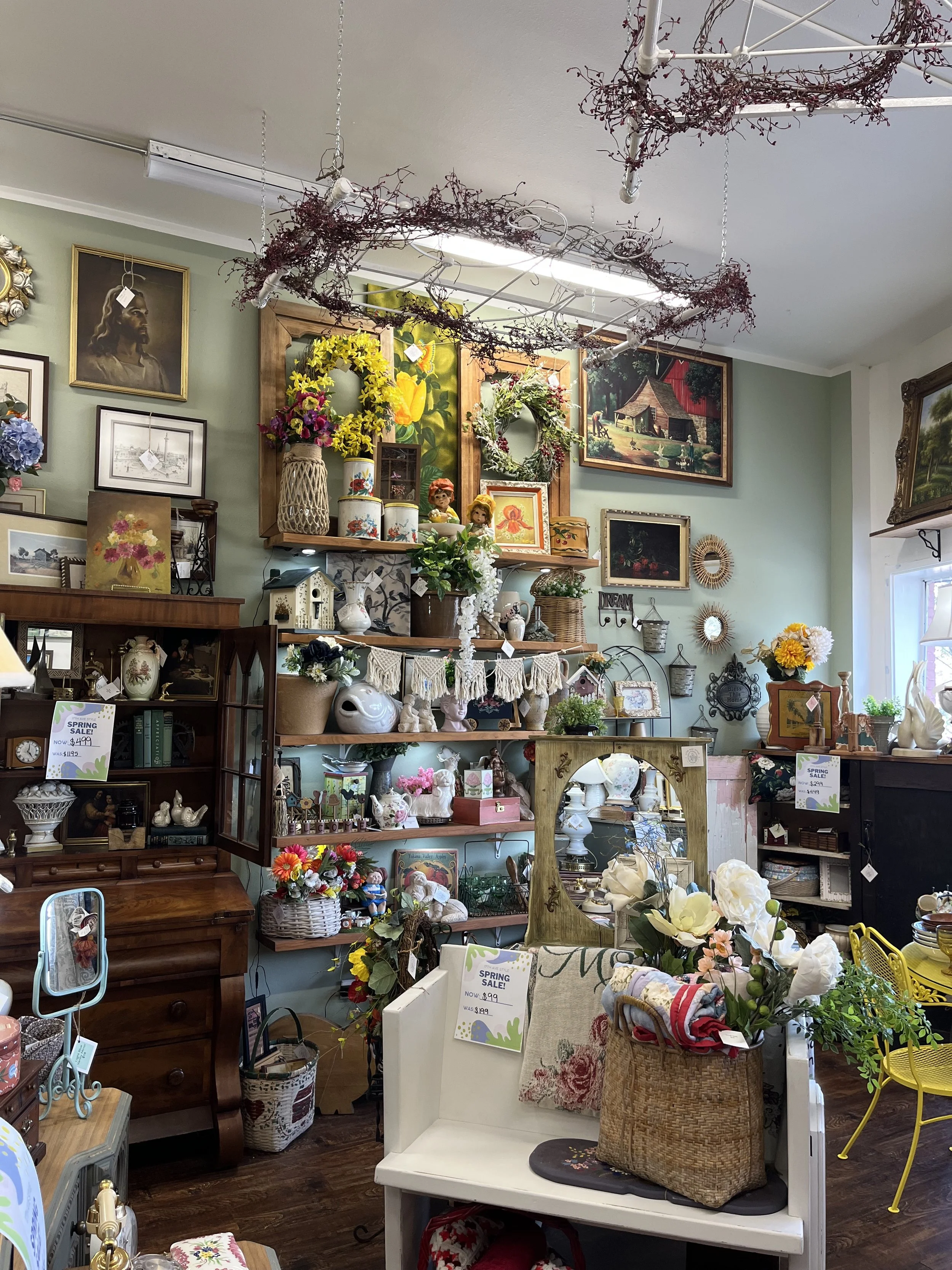 Inside a thrift or vintage store displaying various decorative items, paintings, floral arrangements, and furniture in a cozy, eclectic setup.
