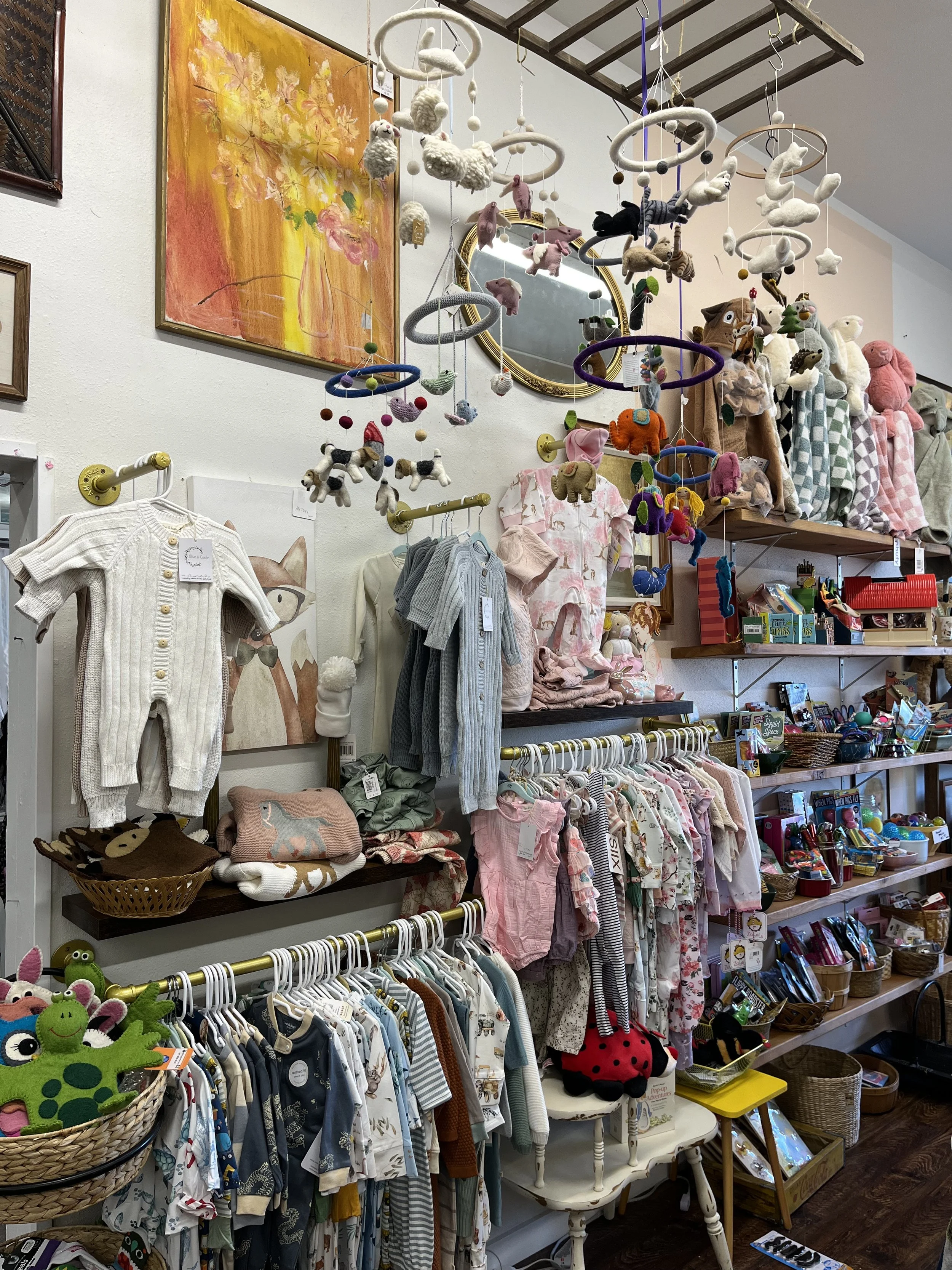 A children's clothing and toy display in a store with hanging stuffed animals, baby clothes on racks, and shelves with various toys and books.
