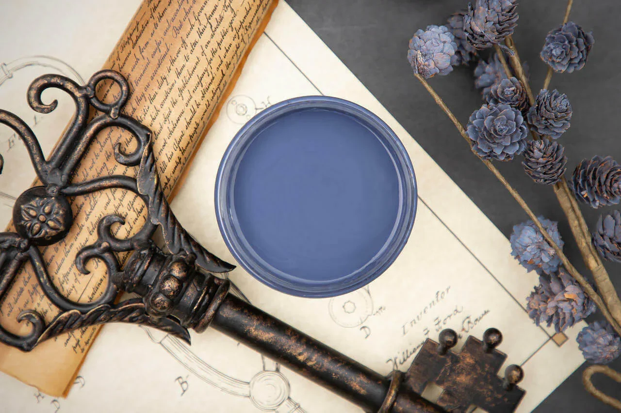 An overhead view of a vintage key resting on architectural blueprints, with a small container of blue paint or liquid and a branch of dried purple flowers nearby.