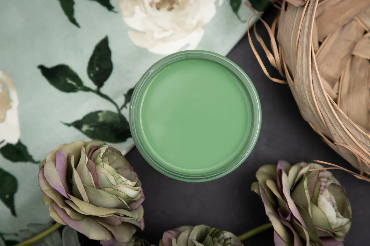 A top view of a green candle surrounded by artificial purple and green roses, a floral patterned cloth, and a woven basket.