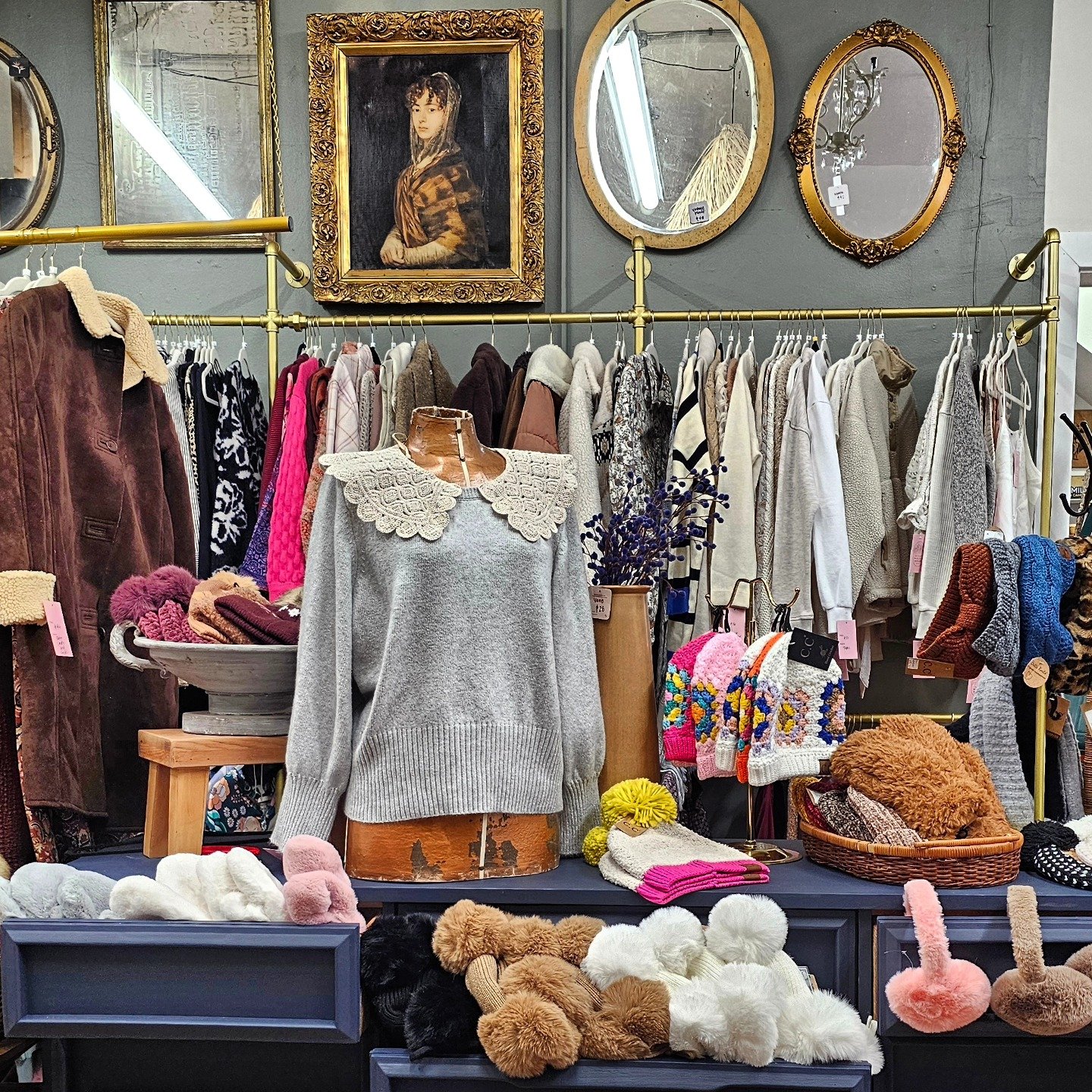 Display of various secondhand clothing, hats, and accessories in a thrift store. There are sweaters, hats, and scarves arranged on tables, with a clothing rack and framed art on the wall above.