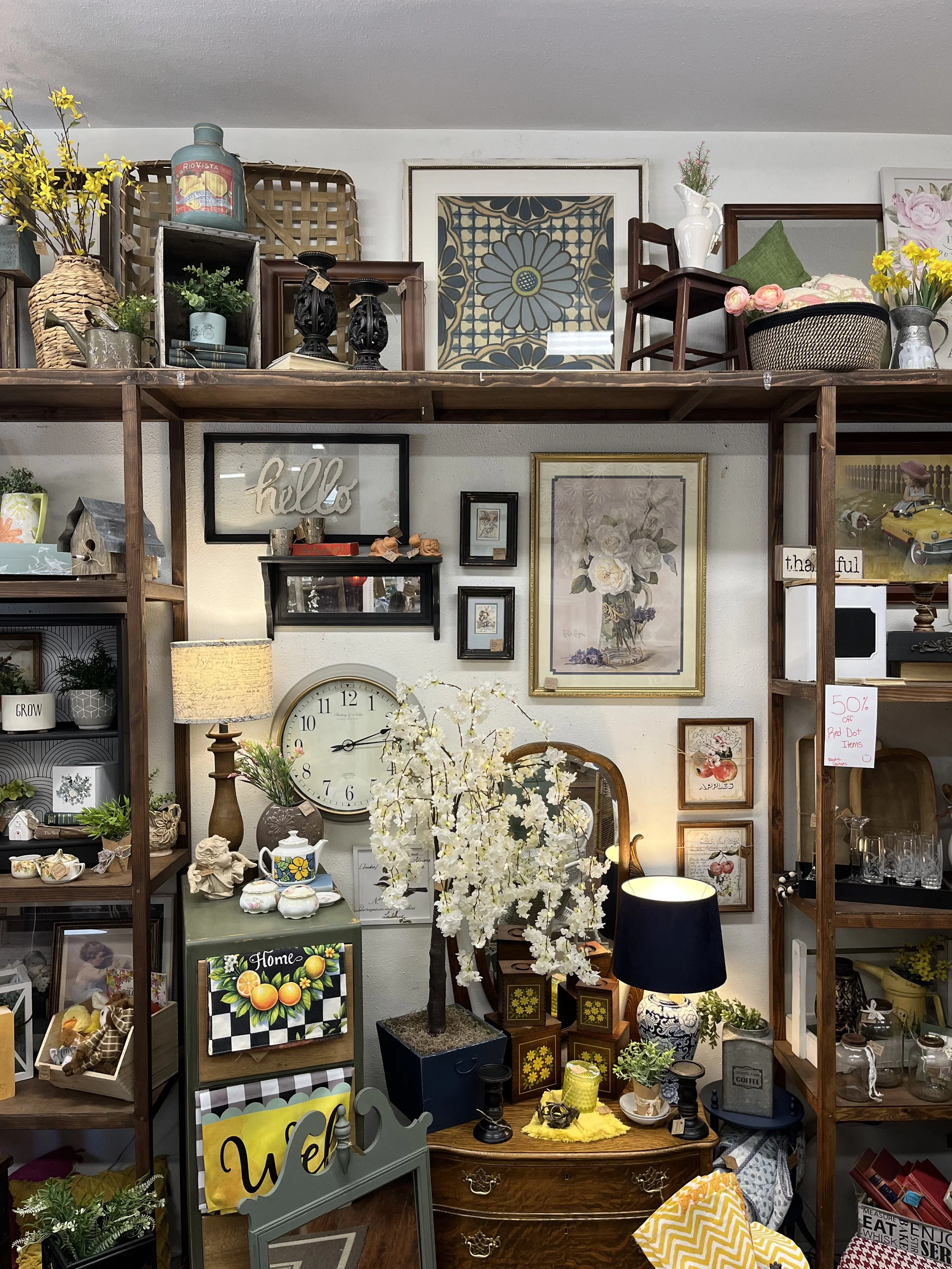Display of various home decor items on shelves and tables, including framed artwork, vases with flowers, a clock, and decorative signs.