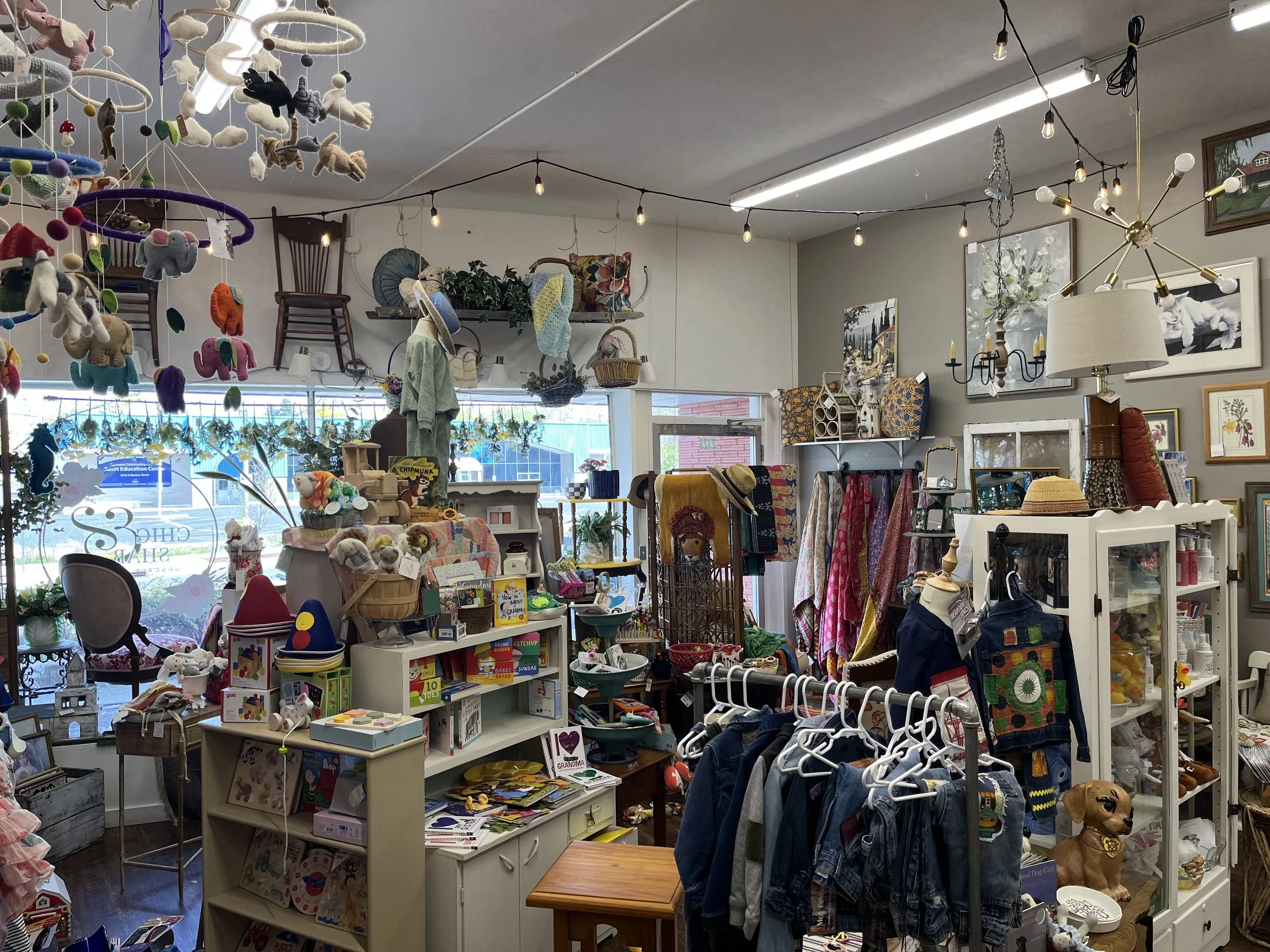 A cluttered retail store with various toys, clothing, and home decor items, including plush toys hanging from the ceiling, framed artwork, lamps, and shelves filled with assorted products.