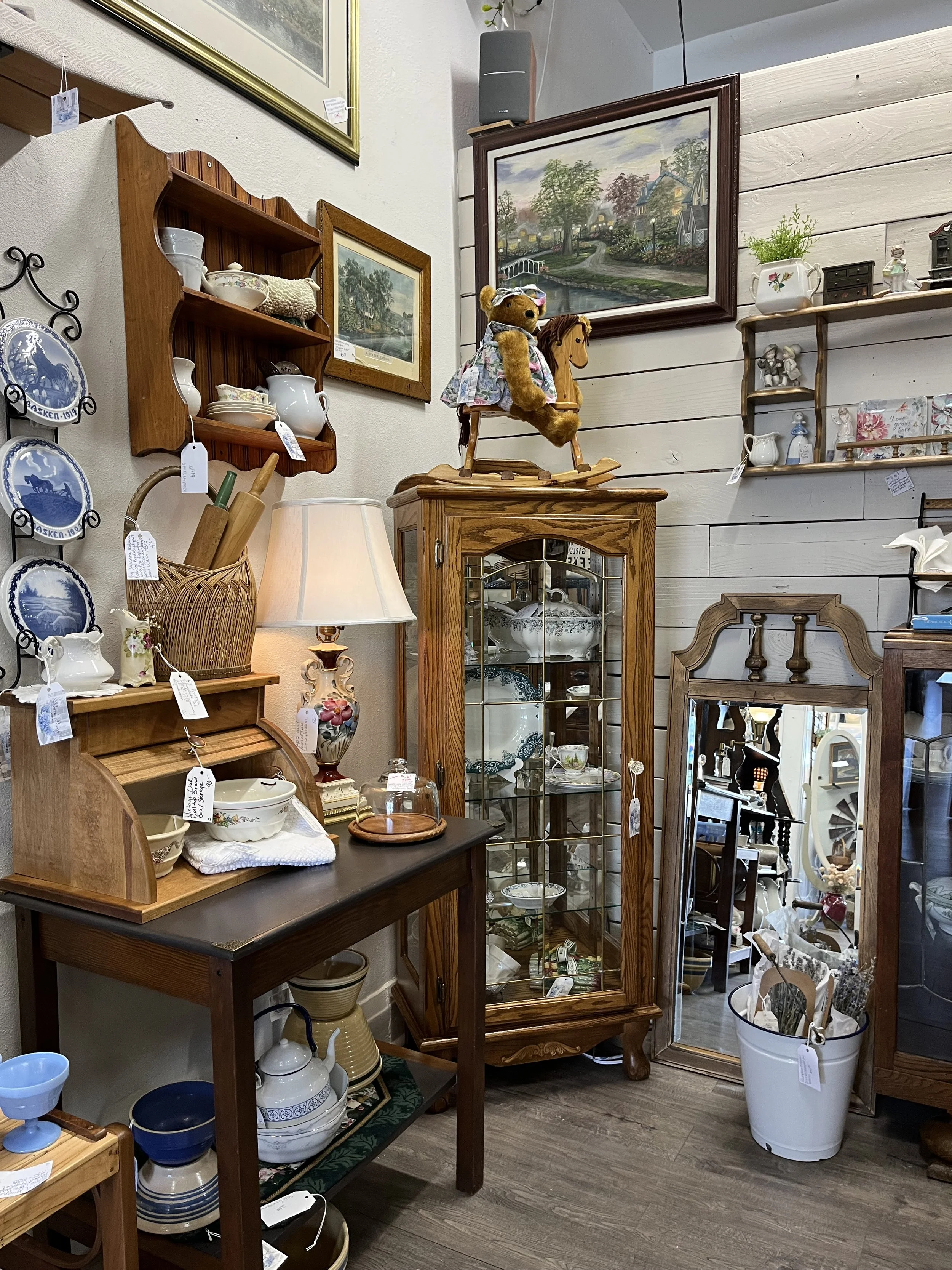 Various antique and vintage ceramic and pottery pieces displayed on shelves and tables in a store, with framed landscape paintings on the wall and a teddy bear on a wooden horse toy.