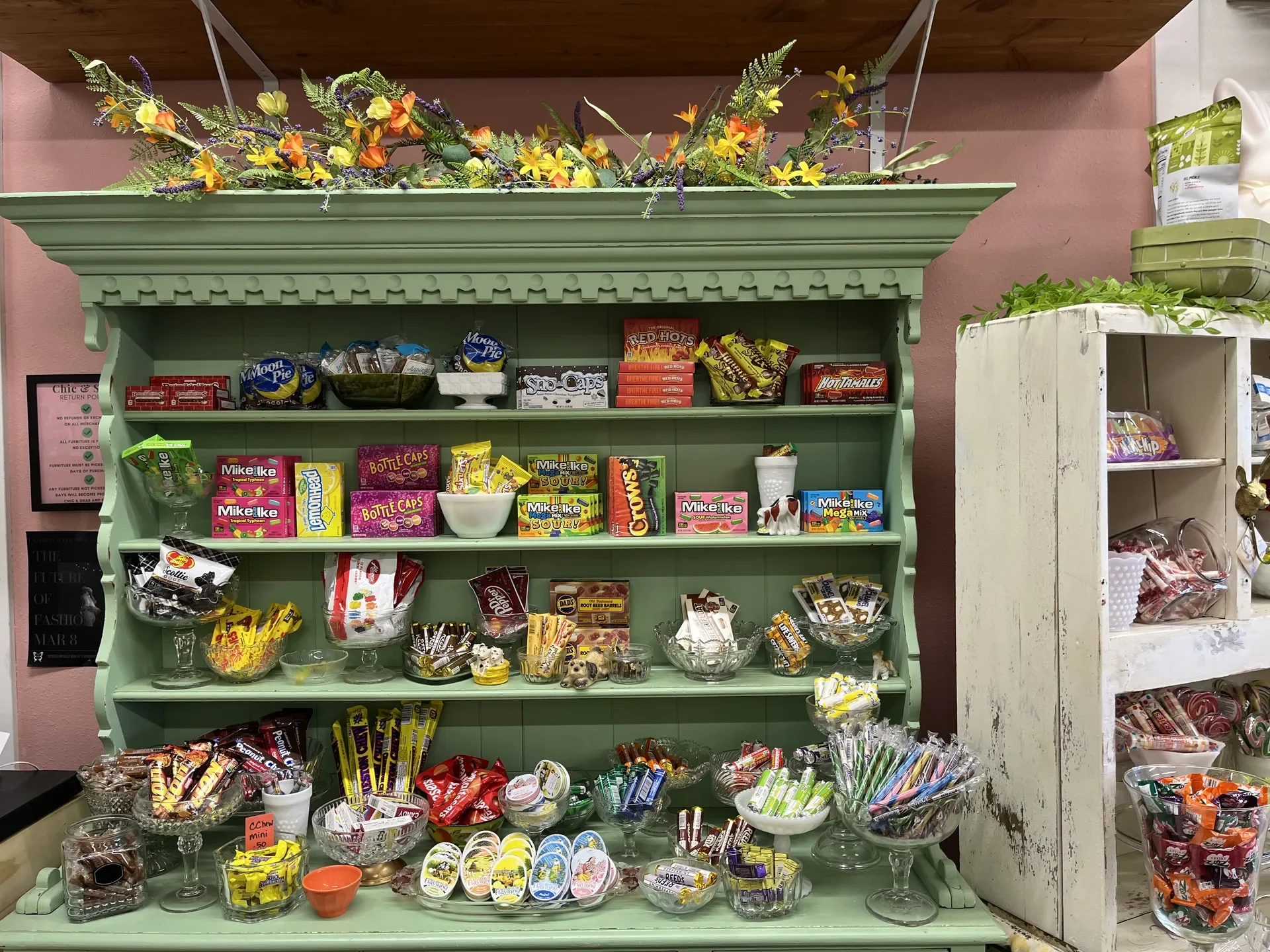 A vintage green candy display shelf filled with various candies and snacks, with decorative flowers on top and pink walls in the background.