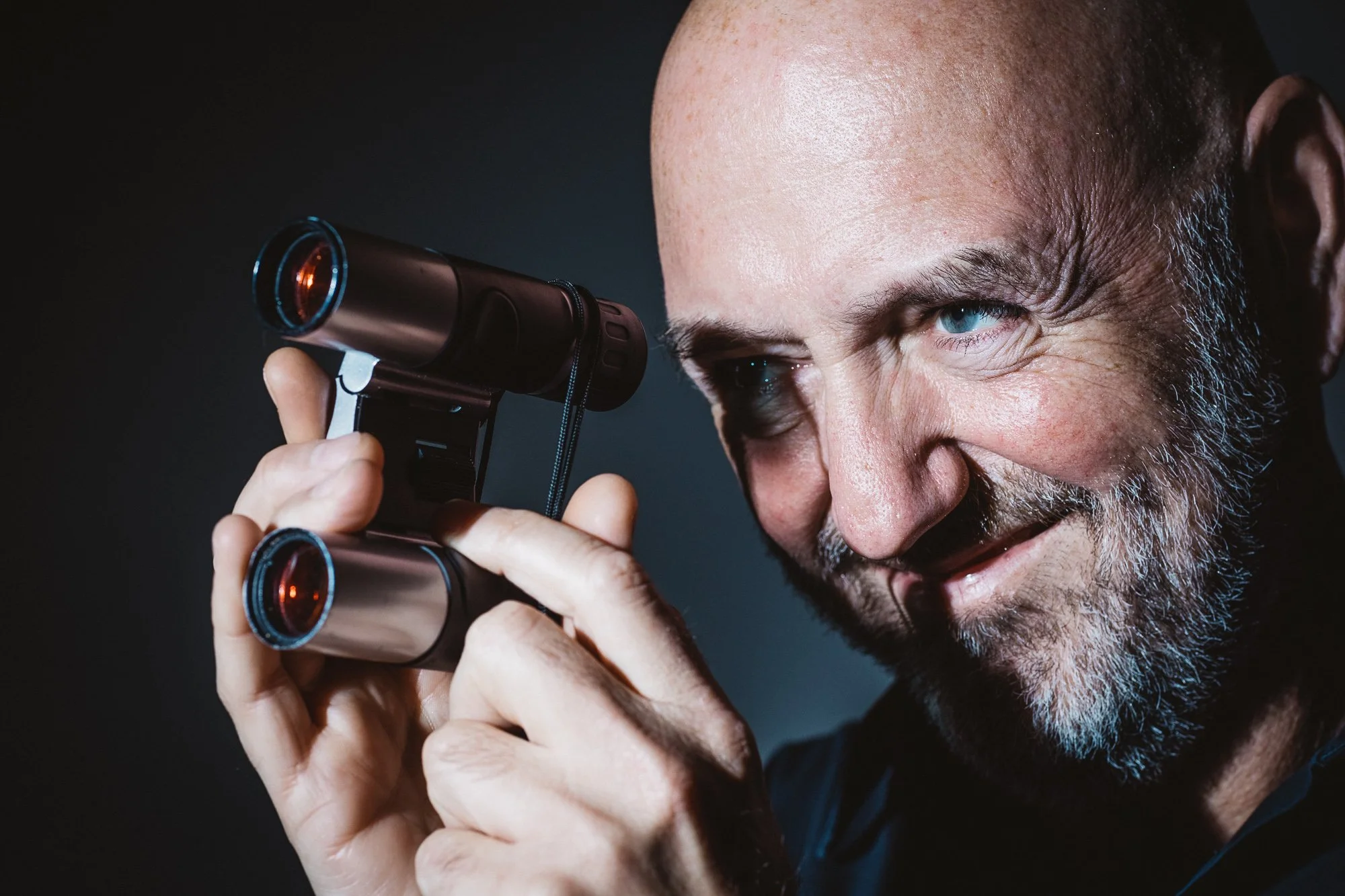 Ein Mann mit blauen Augen hält ein Fernglas ans Auge, im Hintergrund ist ein dunkler Raum.