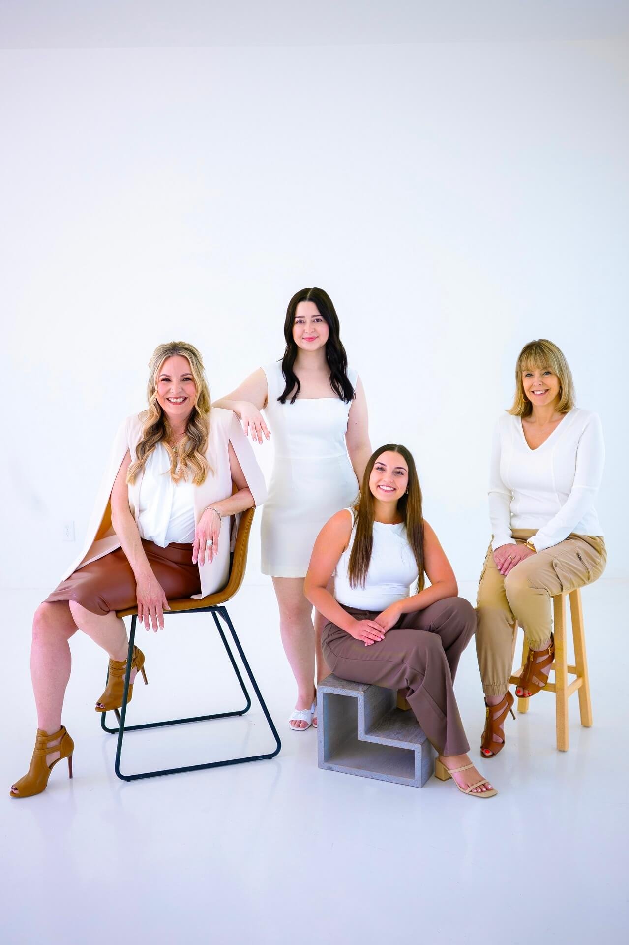 Five women, diverse ages, dressed in white and earth tones, posing in a minimalistic studio with a white background, some sitting and some standing, smiling at the camera.