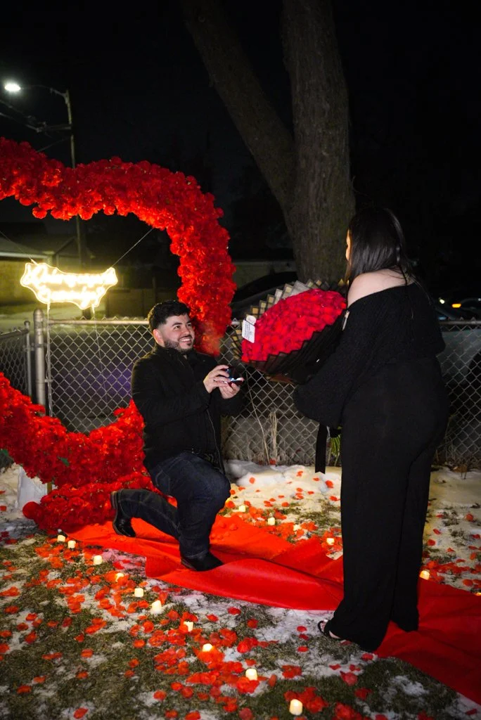 A man proposing marriage on a red carpet in an outdoor setting at night. The man is kneeling with a smile, holding a ring, while a woman stands holding a bouquet of red roses. The scene is decorated with red rose petals and small candles, with a large heart-shaped floral arch in the background.