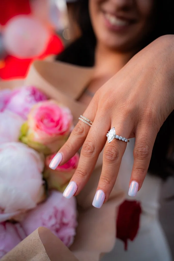 Close-up of a woman's hand displaying an engagement ring with a marquise-shaped diamond, with pink and white flowers in the background.