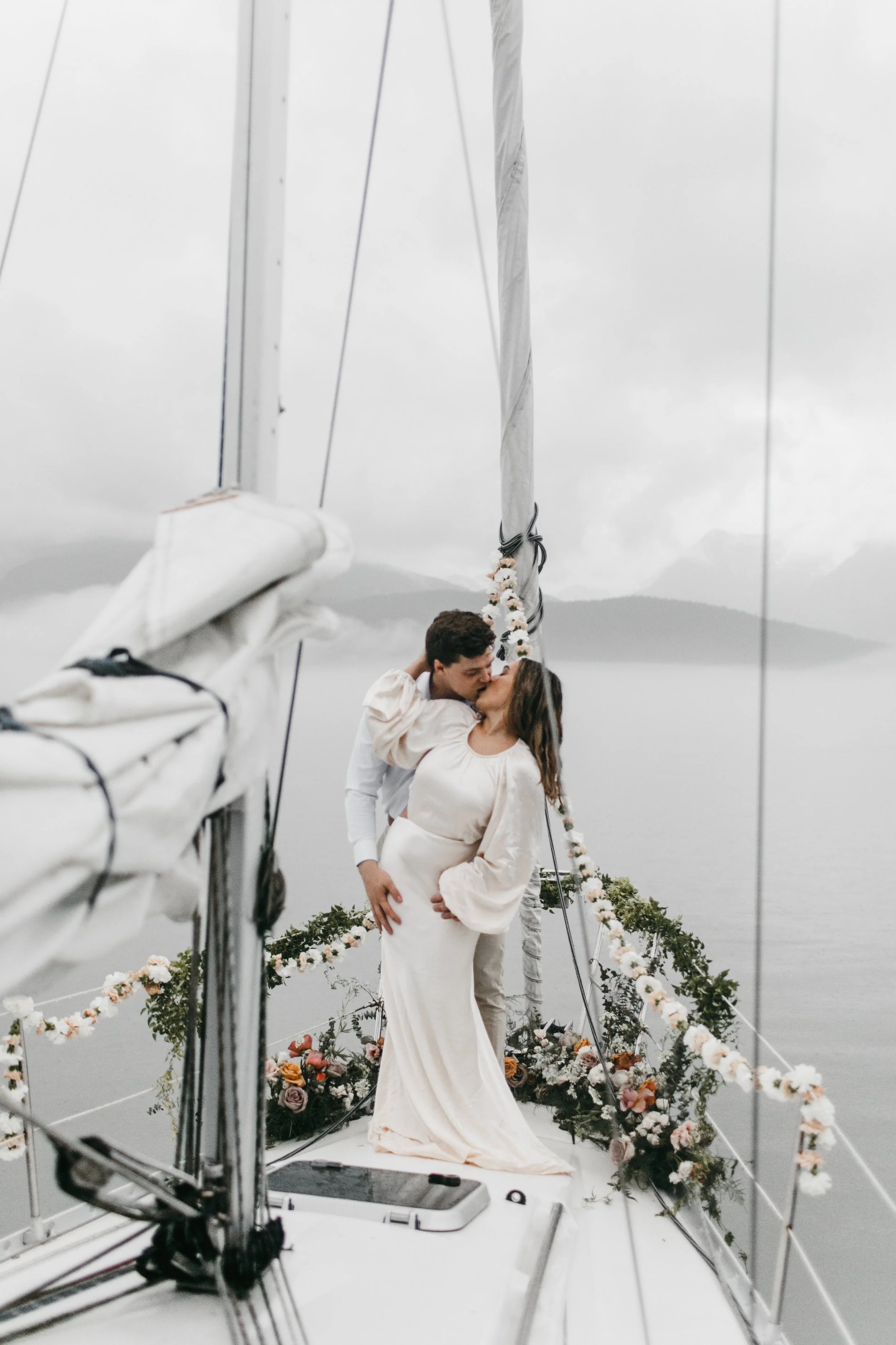 A couple shares a kiss on the deck of a sailboat decorated with flowers and greenery, overlooking a foggy body of water and distant mountains.