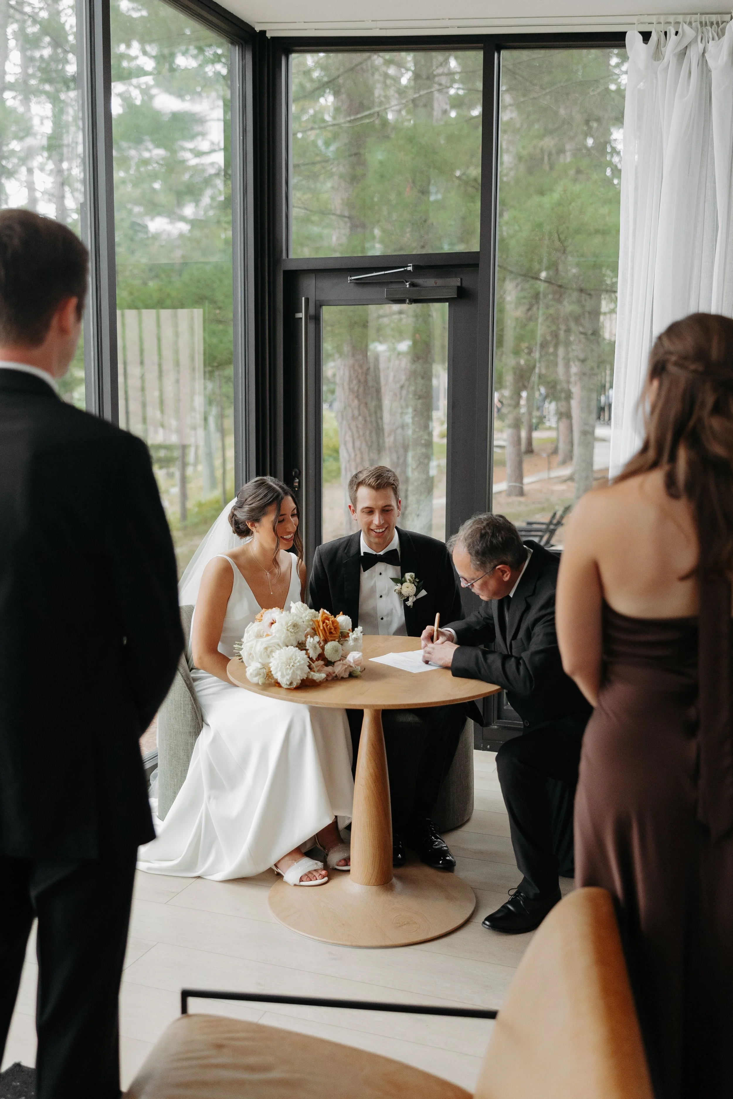 A wedding ceremony with the bride and groom sitting at a small wooden table signing documents, surrounded by friends and family indoors near large windows showing trees outside at Catalyst by Nature Link