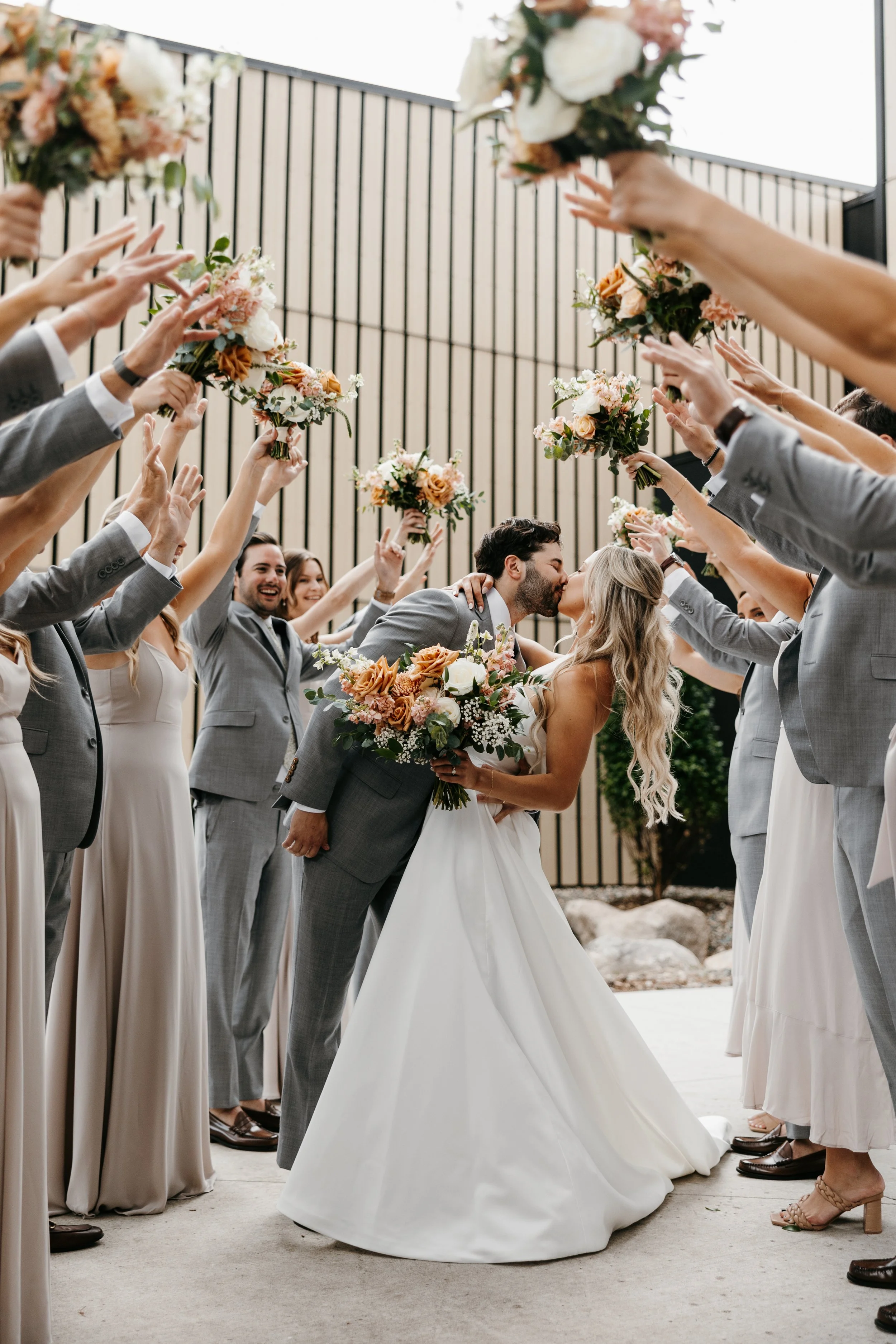 A wedding scene where a bride and groom are kissing under an archway of bridesmaids and groomsmen holding bouquets and raising hands, celebrating.