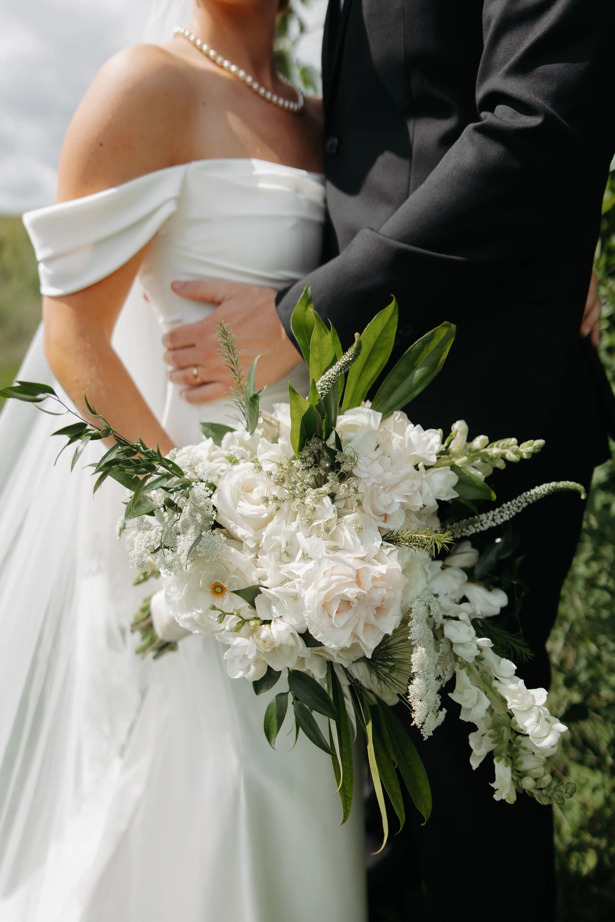 A bride and groom holding a white floral bouquet preside at their wedding, with the bride wearing an off-shoulder white gown and pearl necklace, and the groom in a black suit.