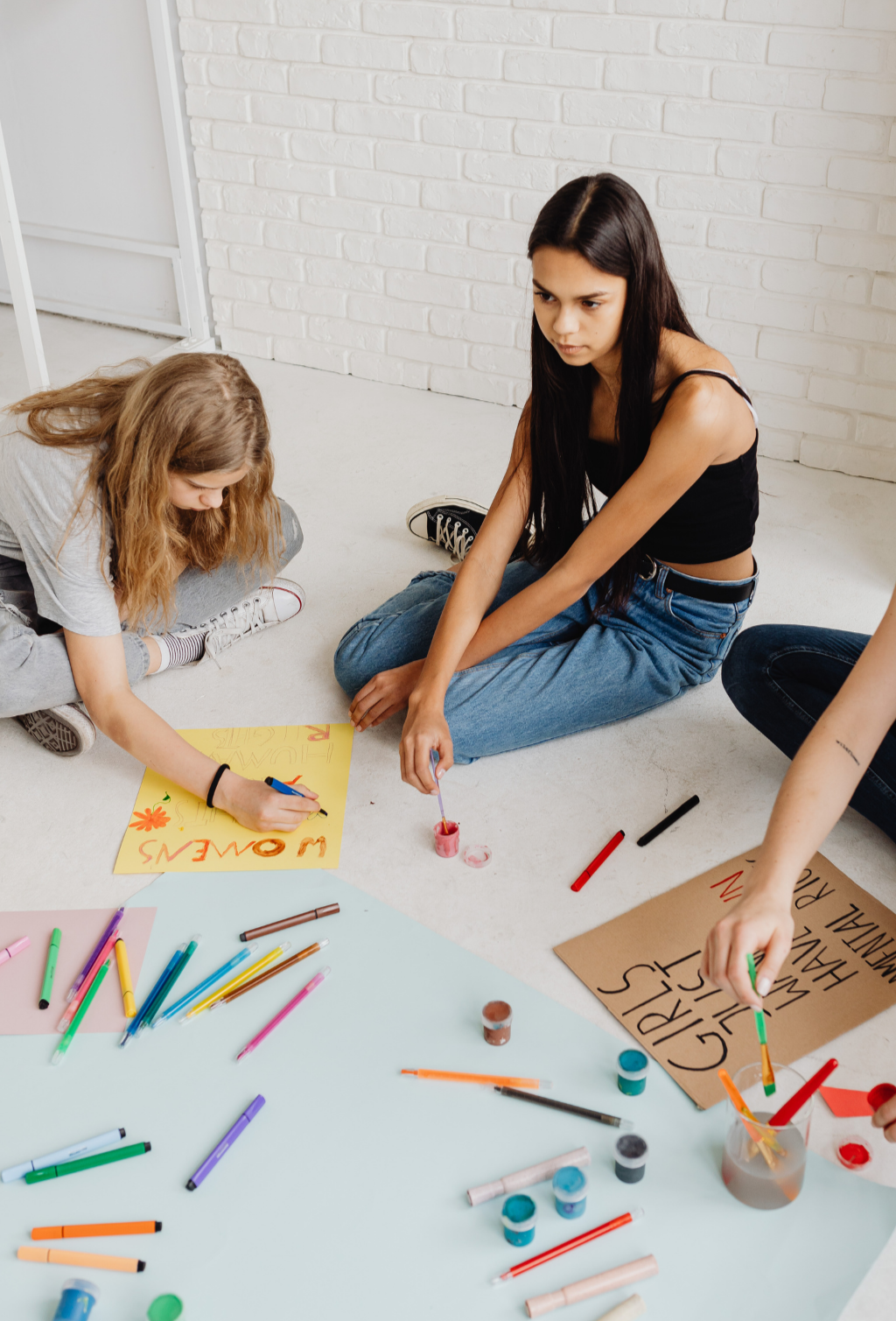 Gruppo di ragazze che creano cartelloni con pennarelli e vernice colorata su un pavimento bianco, decorando con scritte e disegni.