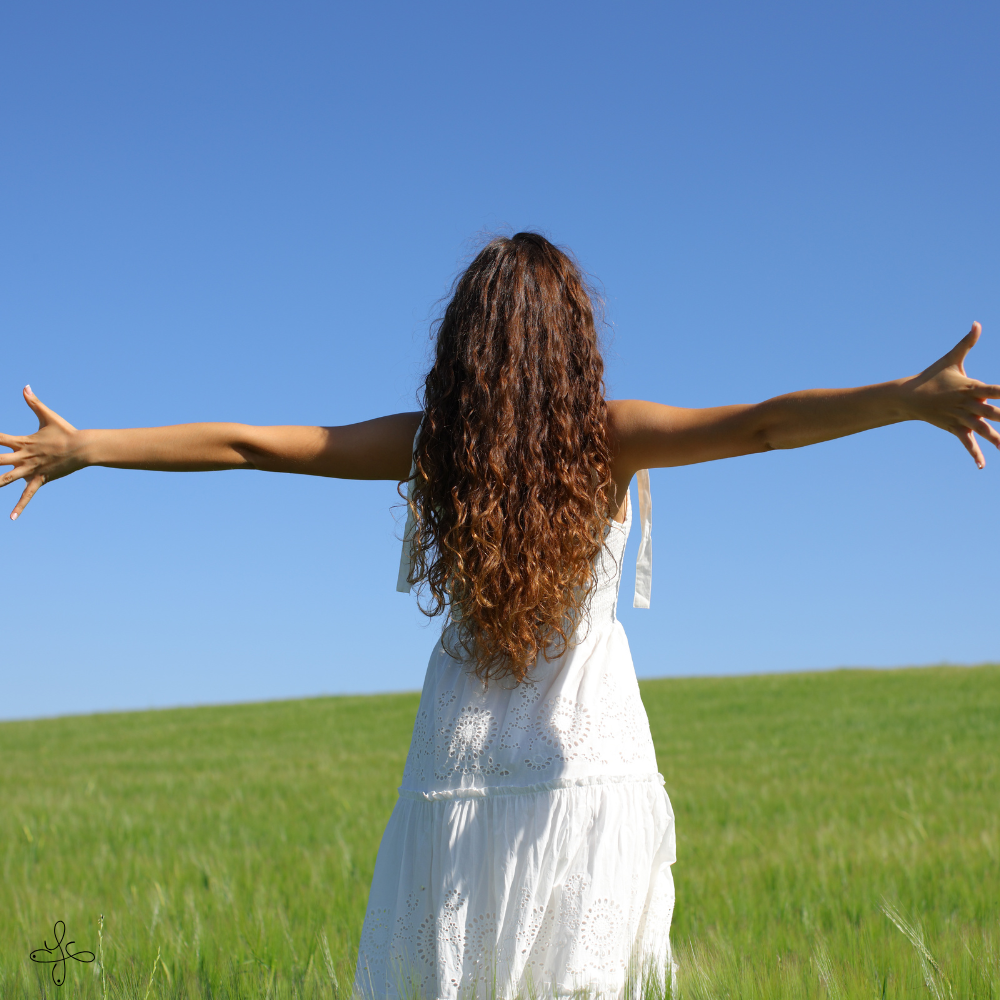Una donna con lunghi capelli ricci indossa un vestito bianco e si sporge in avanti con le braccia aperte in un prato verde sotto un cielo azzurro.